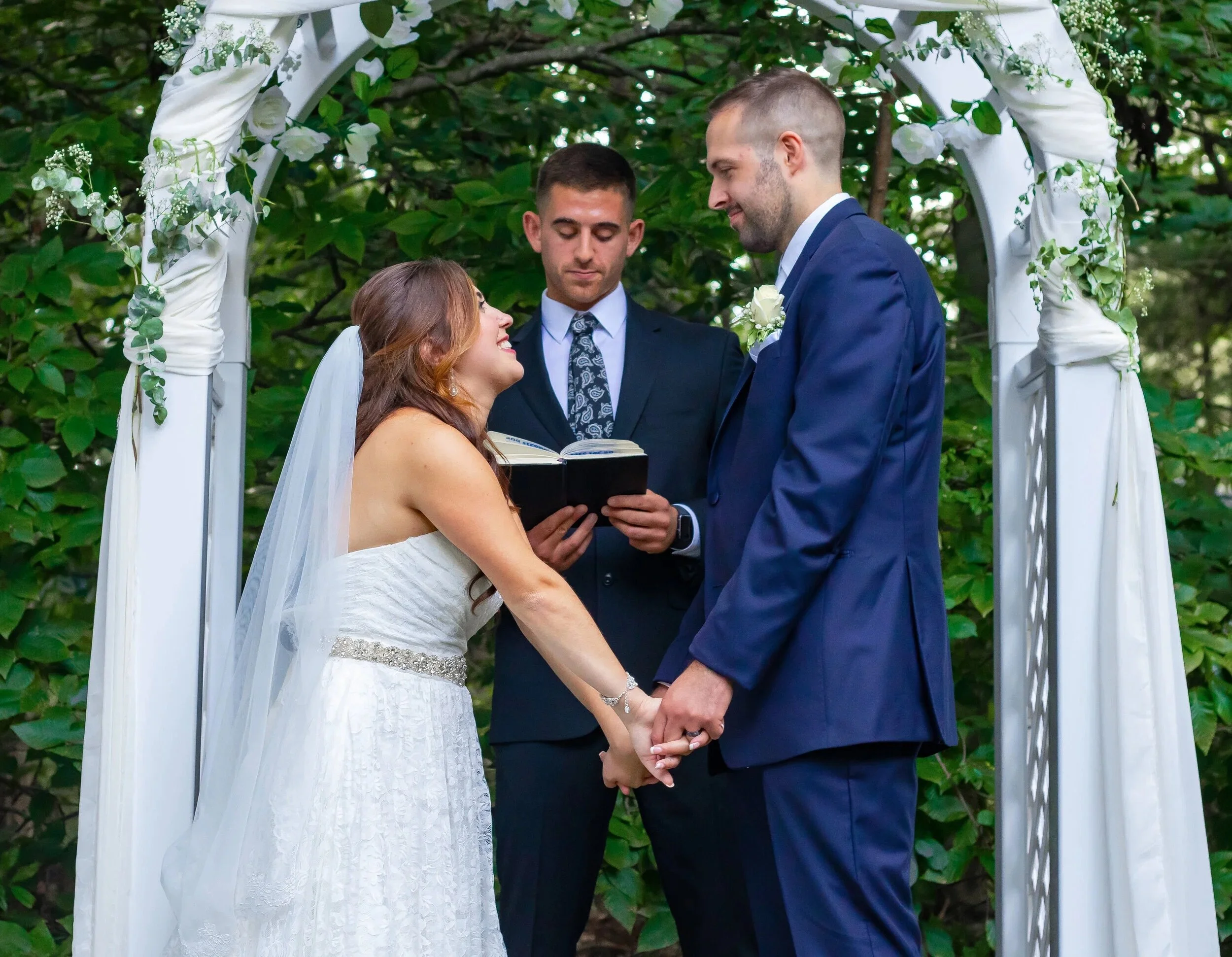 Couple holding hands, getting married outdoors under a white floral arch with an officiant standing behind them
