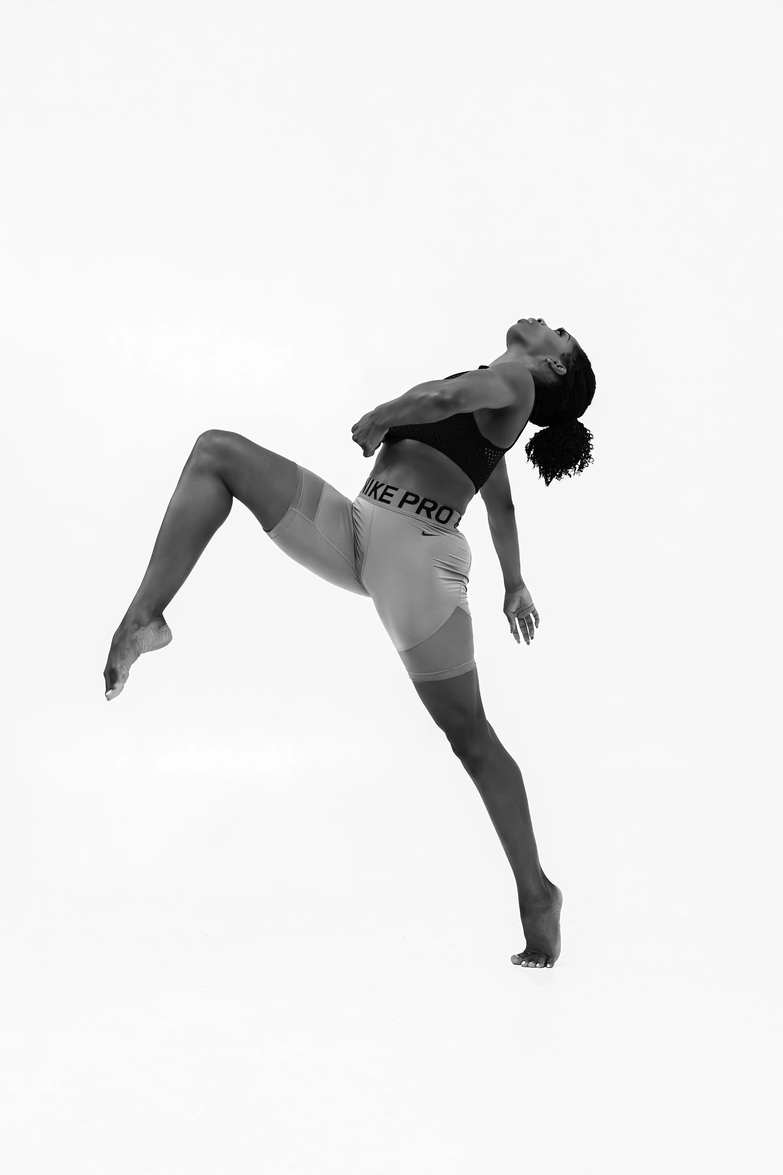 A black and white photograph of a woman performing a yoga pose on a plain white background. She is balancing on one foot with her arms crossed over her chest, and her head tilted back with eyes closed.