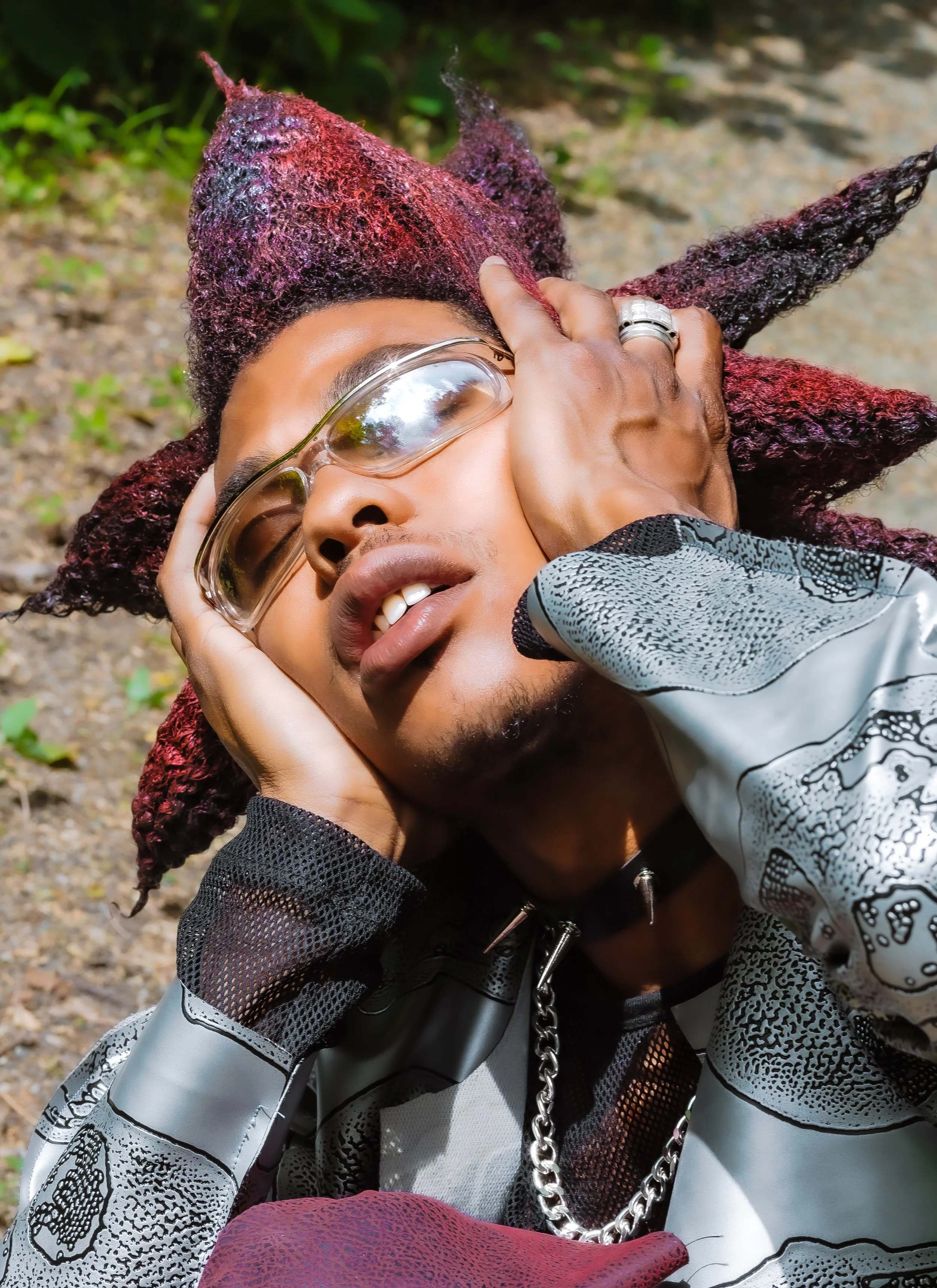 A person with dark skin, purple and black textured hair, wearing protective glasses, a patterned jacket, and a chain necklace, is lying on the ground outdoors with their eyes closed, holding their head with both hands, and appearing to be in discomfo
