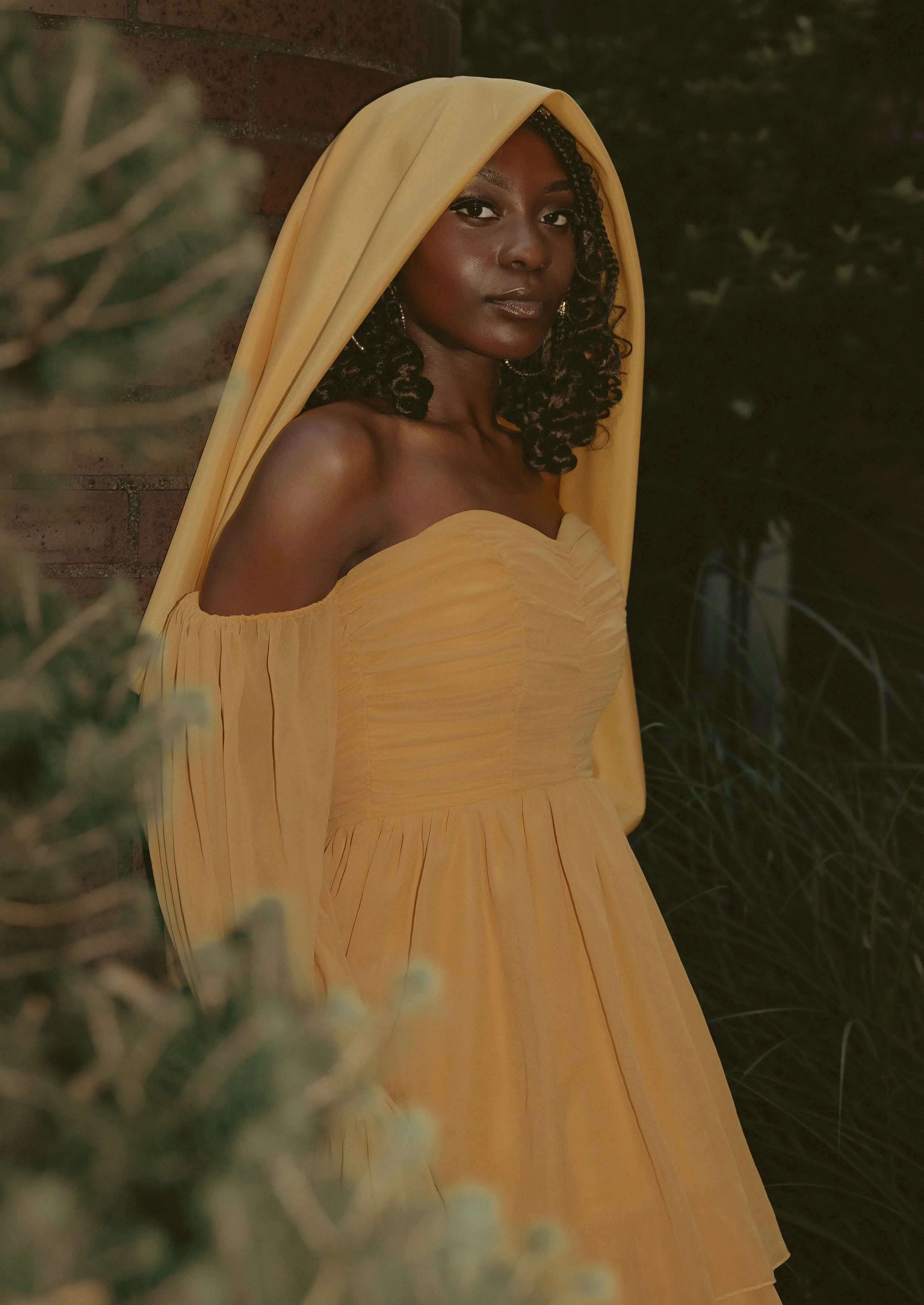 A woman with dark skin, curly hair, and a serious expression, wearing a yellow off-shoulder dress with a matching yellow hood, standing outdoors near a brick wall and greenery.
