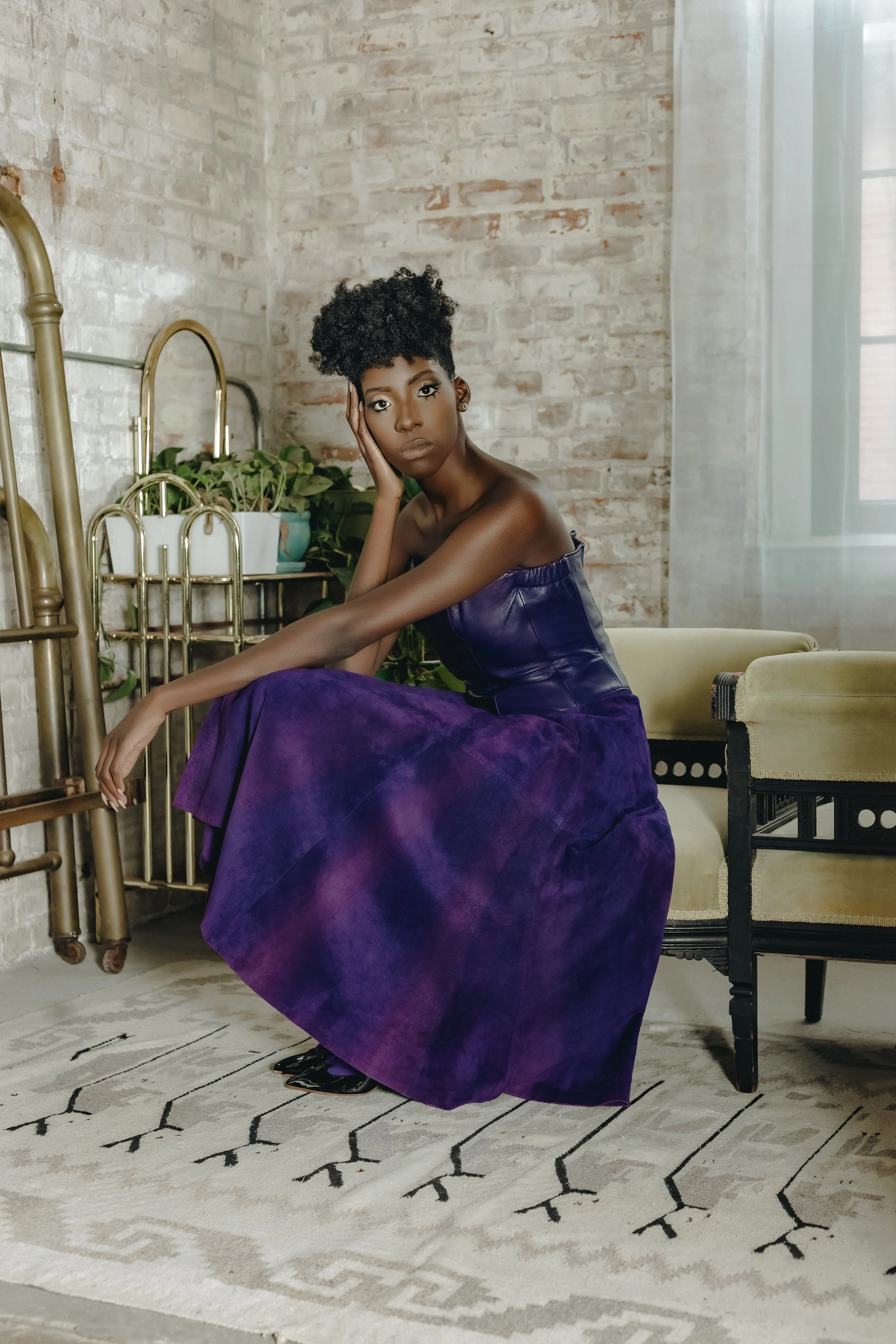 A woman with short curly hair, wearing a purple strapless dress, sitting on a beige and black vintage sofa in a room with exposed brick wall, green plants, and a patterned rug.