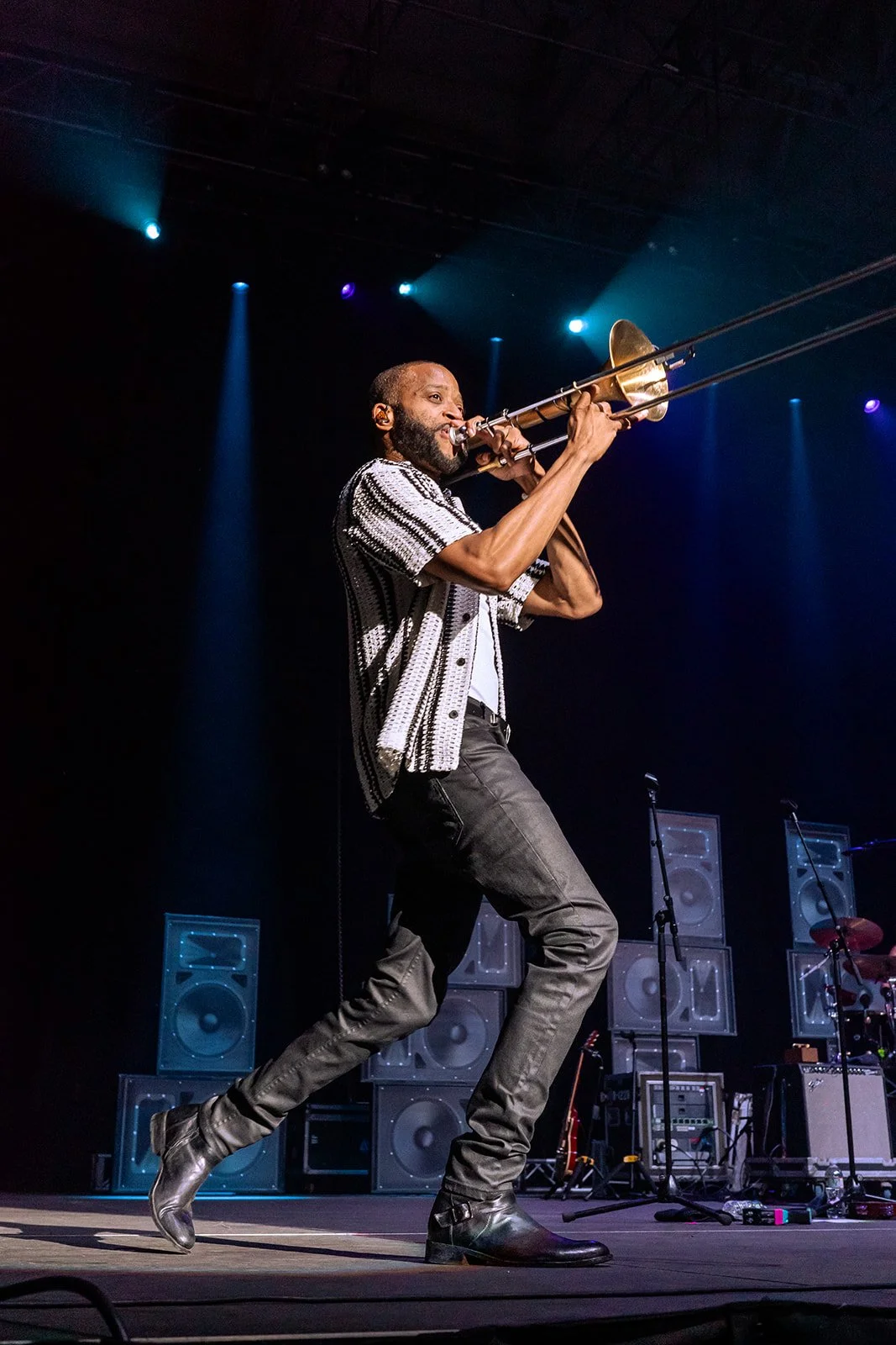 A man playing a trombone on stage during a concert, with large speakers behind him and stage lighting overhead.