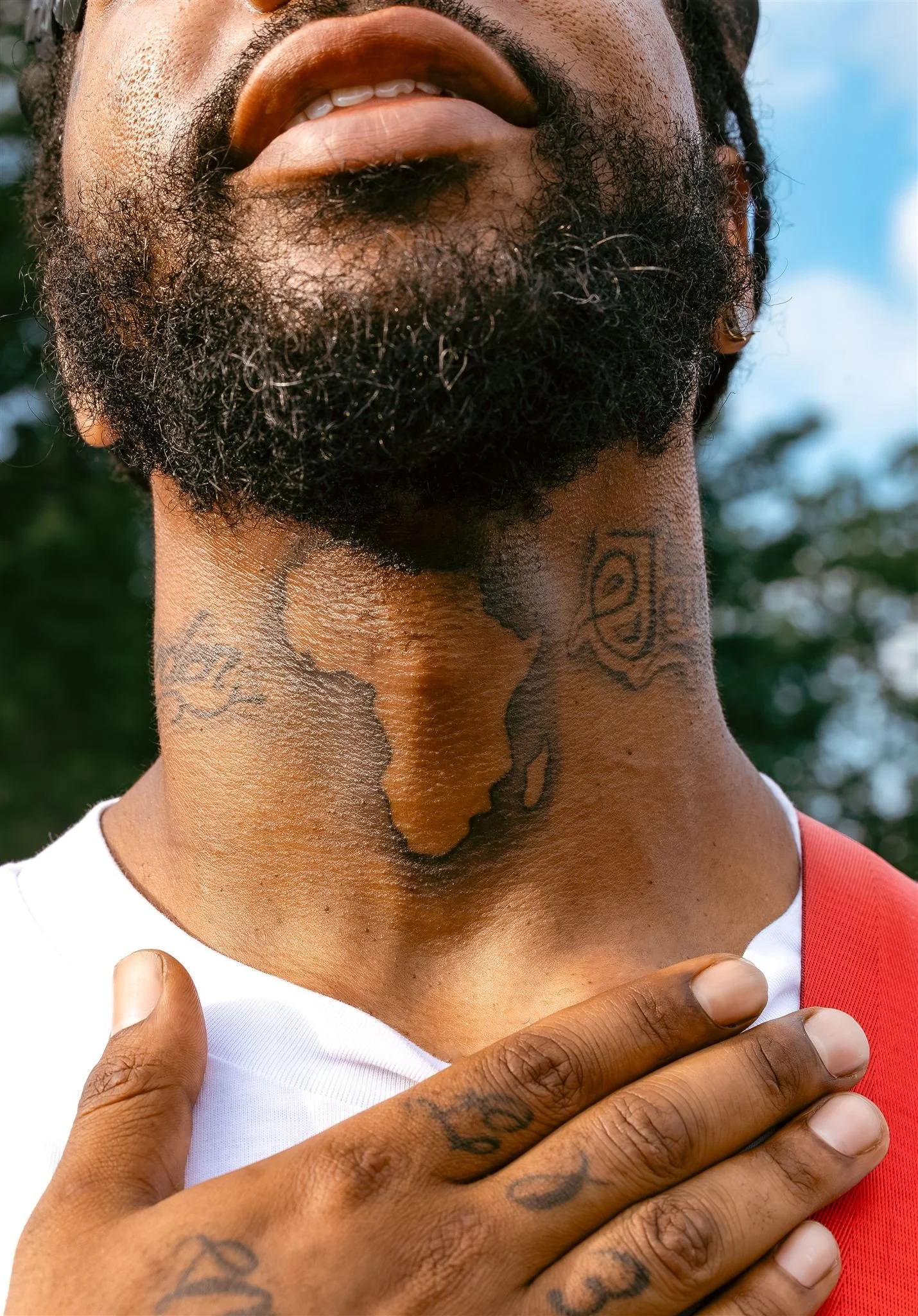 Close-up of a man with a beard flipping his head back slightly, showing tattoos on his neck and hand, with one hand over his chest. The background shows blurred green trees and a blue sky.