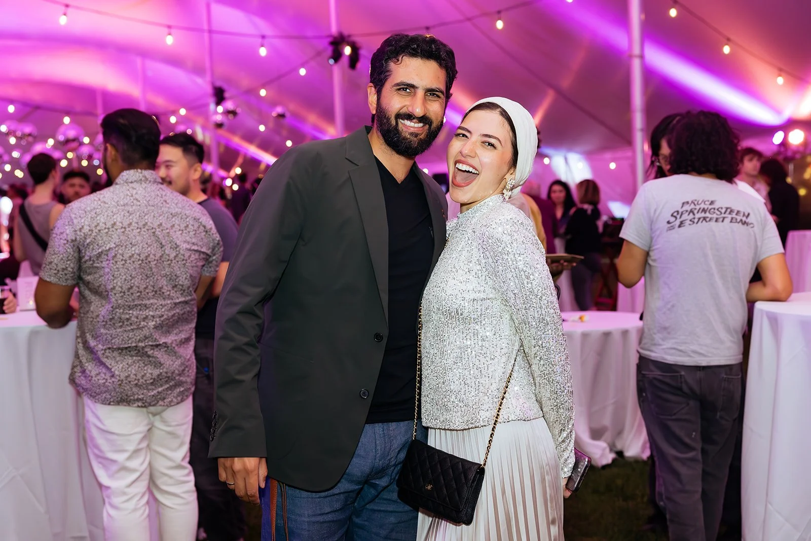 A man and woman smiling and posing together at a lively party or event inside a tent with pink and purple lighting. The woman is wearing a white headscarf and a shiny silver jacket, and the man is in a dark blazer. They are surrounded by other guests