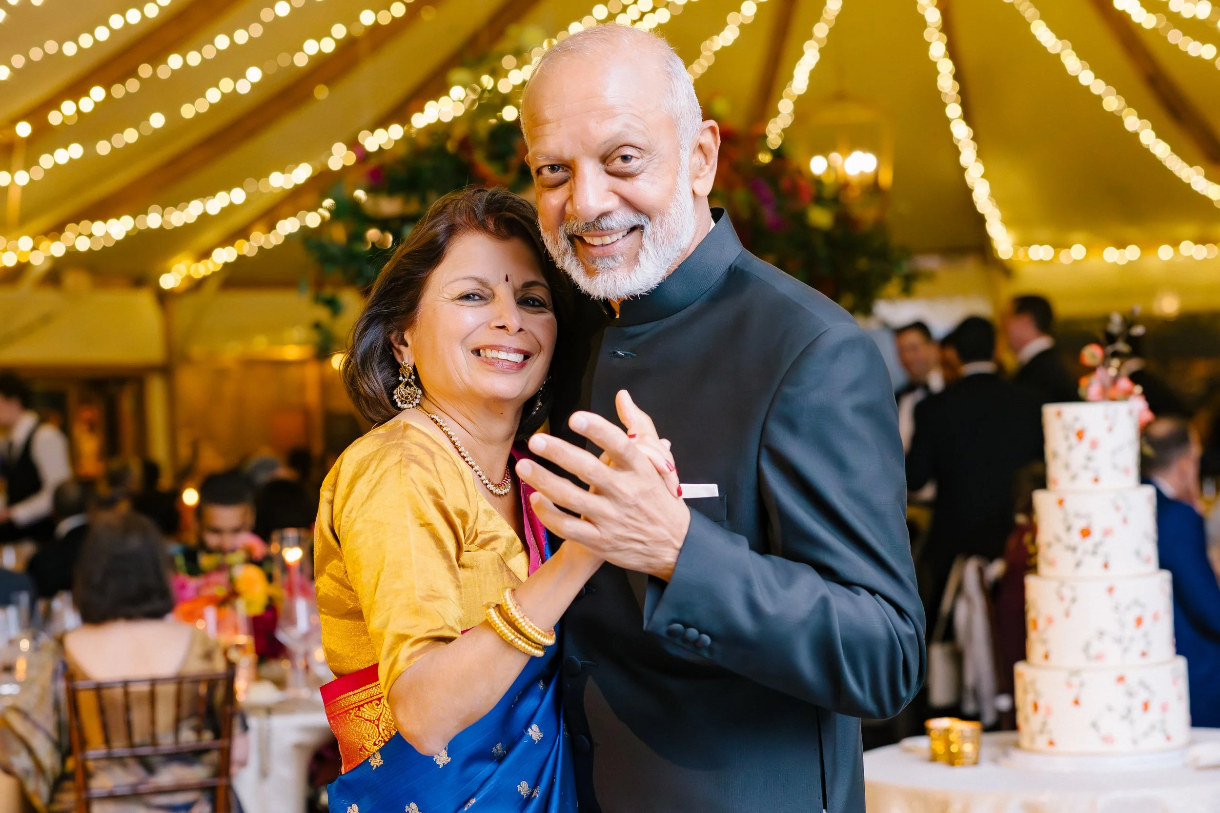 An elderly couple dancing at a wedding reception, surrounded by string lights, a multi-tiered wedding cake, and guests in the background.