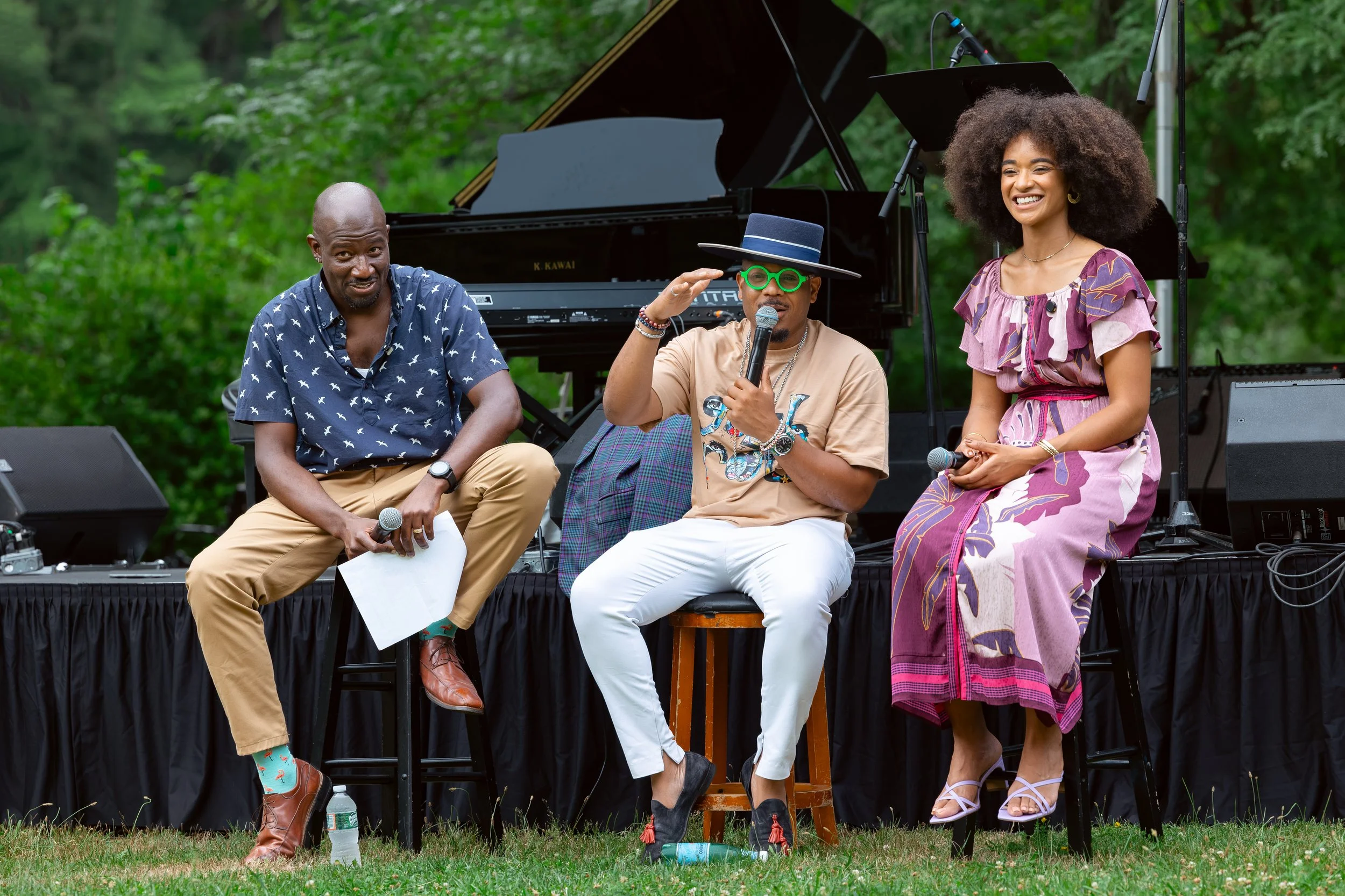 Four people sitting on a stage outdoors with a grand piano in the background. They are engaged in a discussion or performance, smiling and holding microphones. The person on the left is wearing a blue patterned shirt and beige pants, the second perso