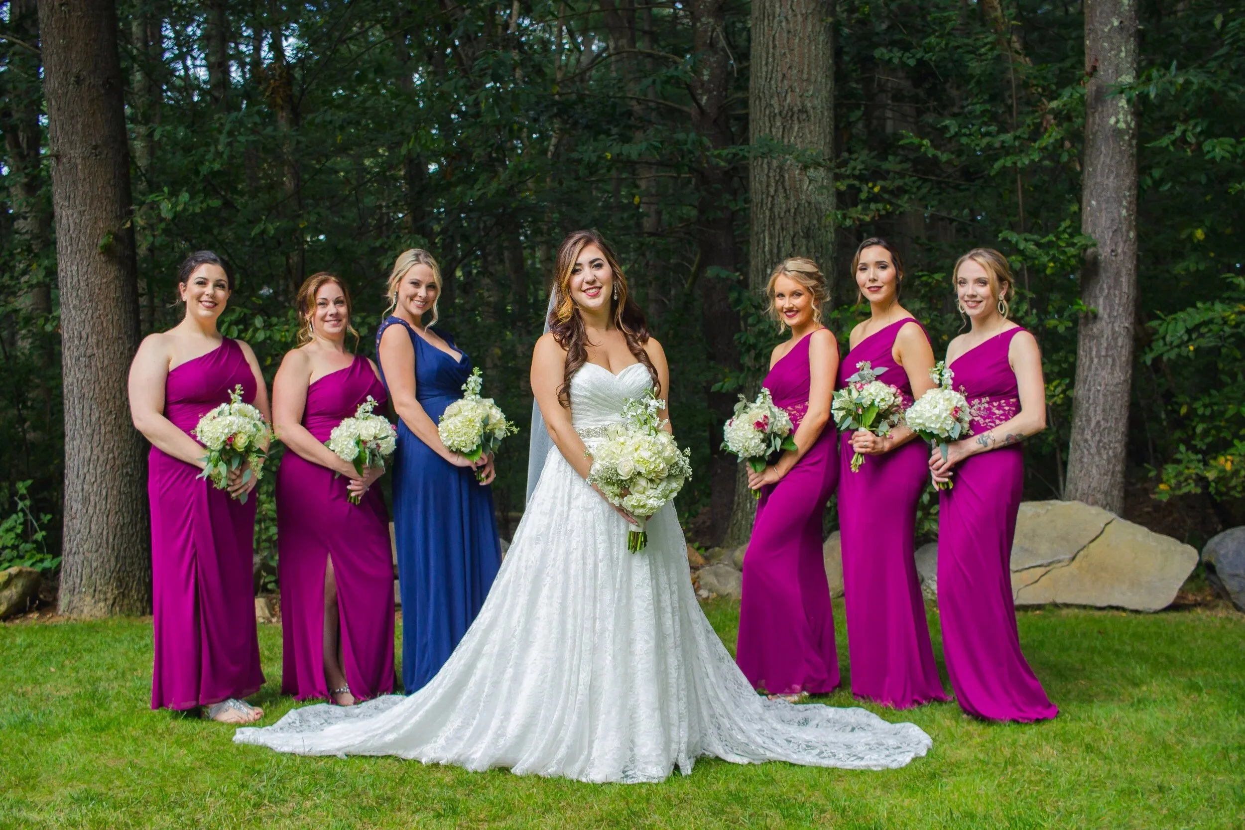 A bride in a white wedding dress holding a bouquet stands among six bridesmaids in purple and blue dresses, all holding bouquets, outdoors in a wooded area.