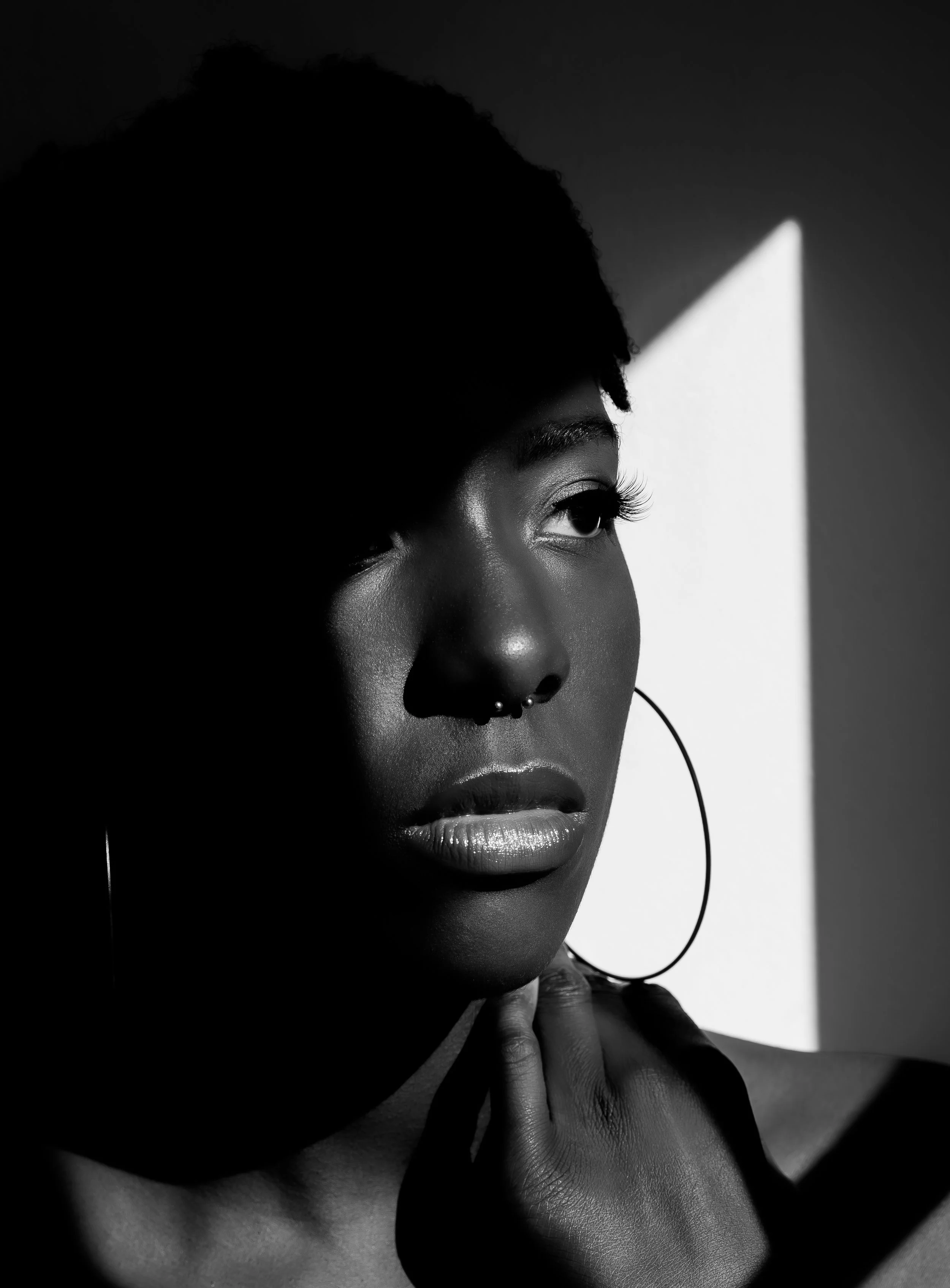 Black and white portrait of a woman with short curly hair, large hoop earrings, and a septum piercing, with shadows and light creating a dramatic effect.