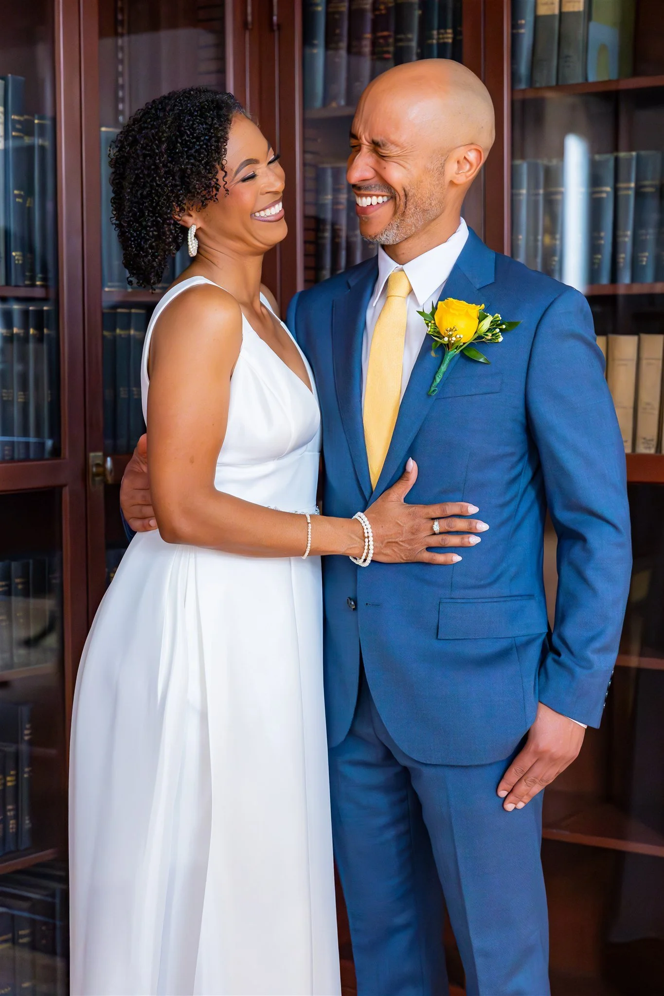 A happy bride and groom smile and hug each other in front of a bookshelf, dressed in wedding attire with the bride in a white gown and the groom in a blue suit with a yellow tie and boutonniere.