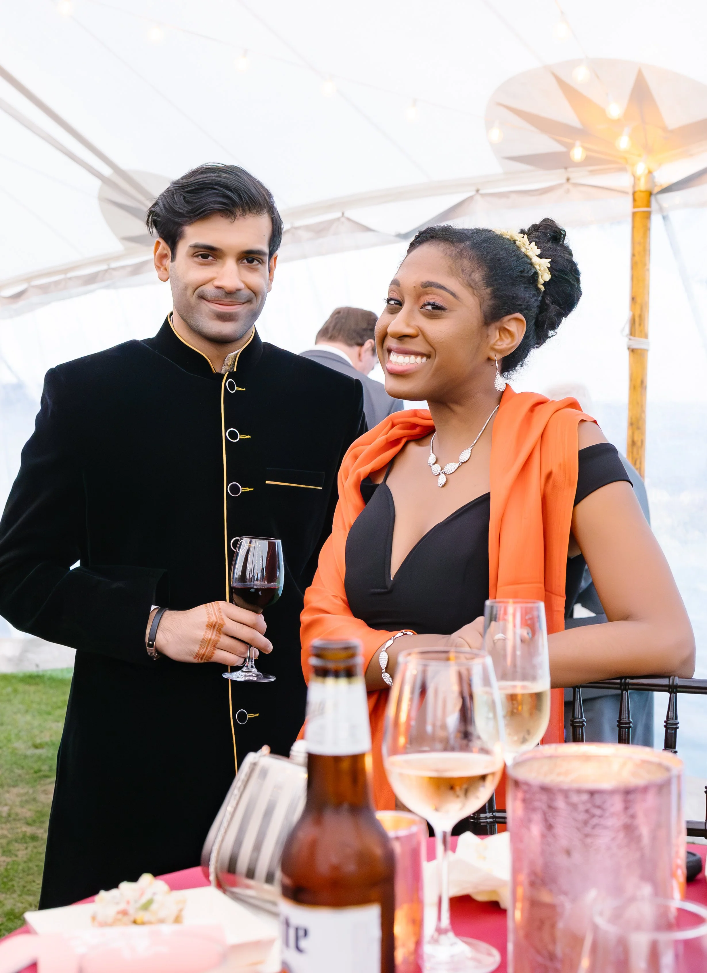 A man in a black traditional suit and a woman in a black dress with an orange shawl, smiling at a social event with drinks on the table.