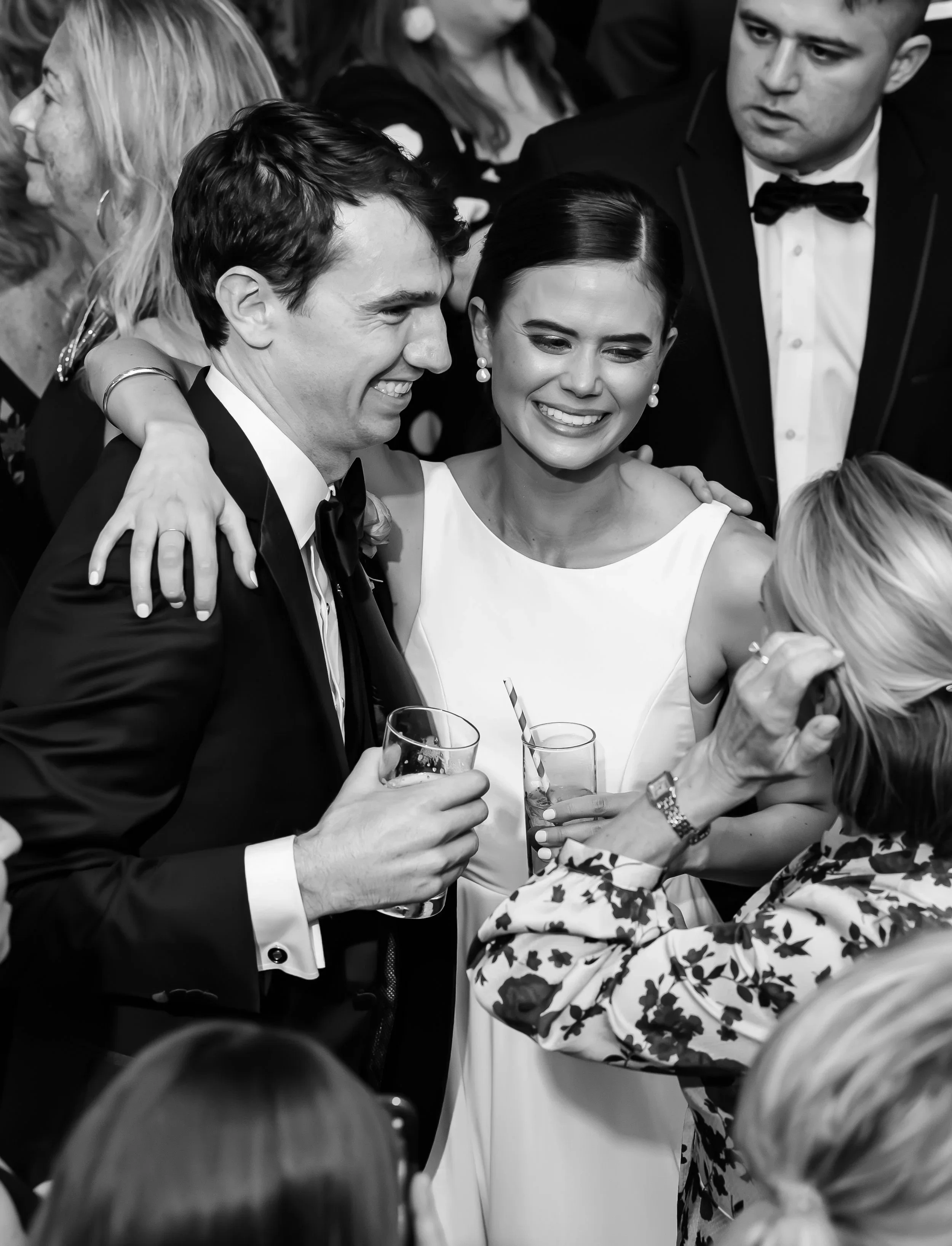 Black and white photo of a celebration with smiling people, including a woman in a white dress and a man in a tuxedo, holding drinks, surrounded by others in formal attire.