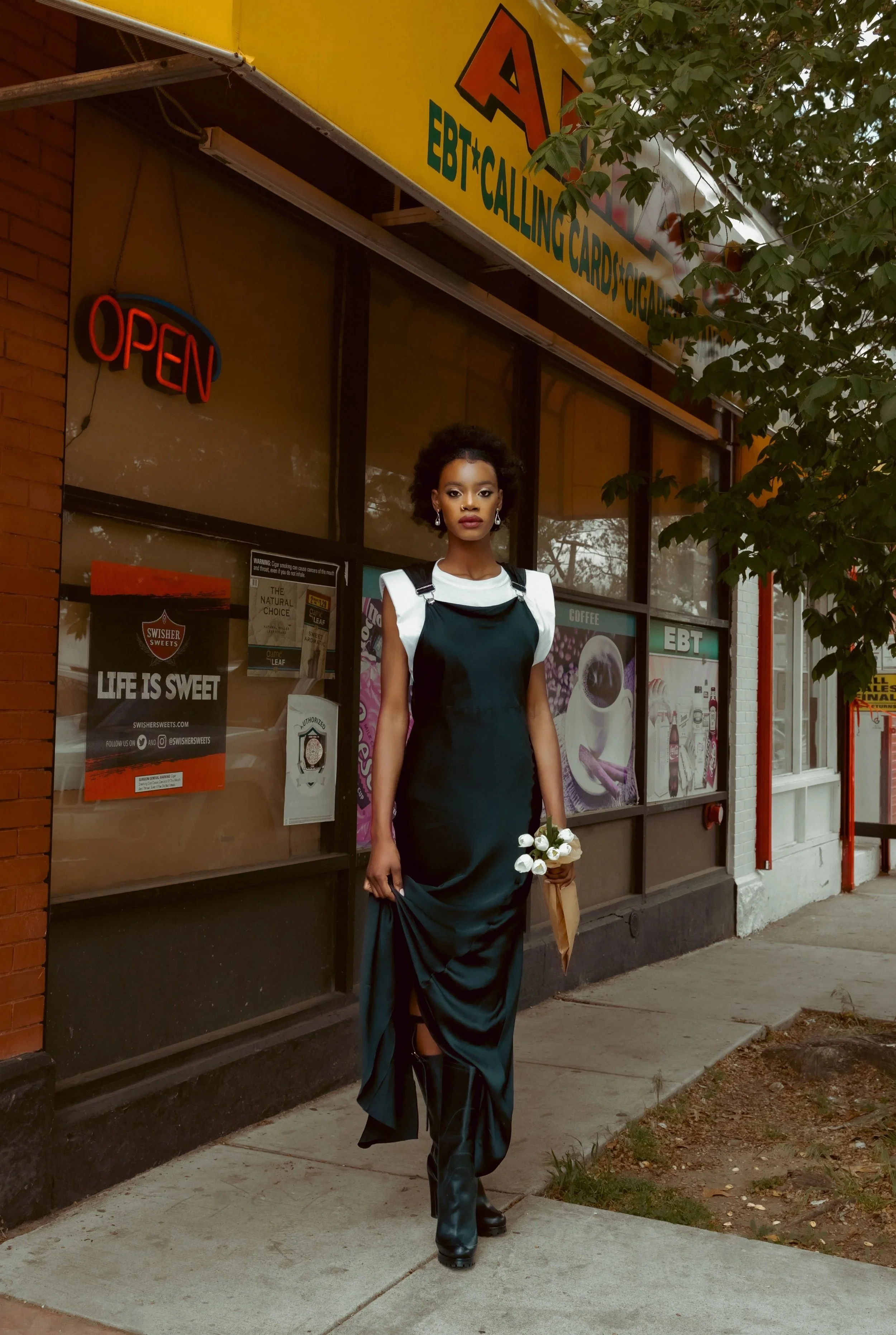 A woman standing on a sidewalk in front of a store with a yellow sign, holding a bouquet of white roses, wearing a black dress over a white shirt with puffed sleeves, and black knee-high boots.