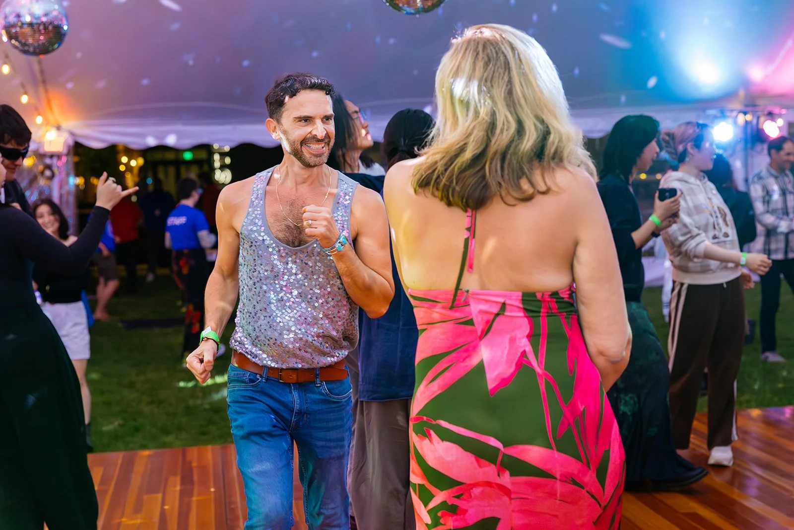 A man and a woman dance and smile at a party under a white tent with disco balls and colorful lights. The man is wearing a sparkly tank top and jeans, while the woman wears a bright pink and green floral dress with bare shoulders. Other people are da
