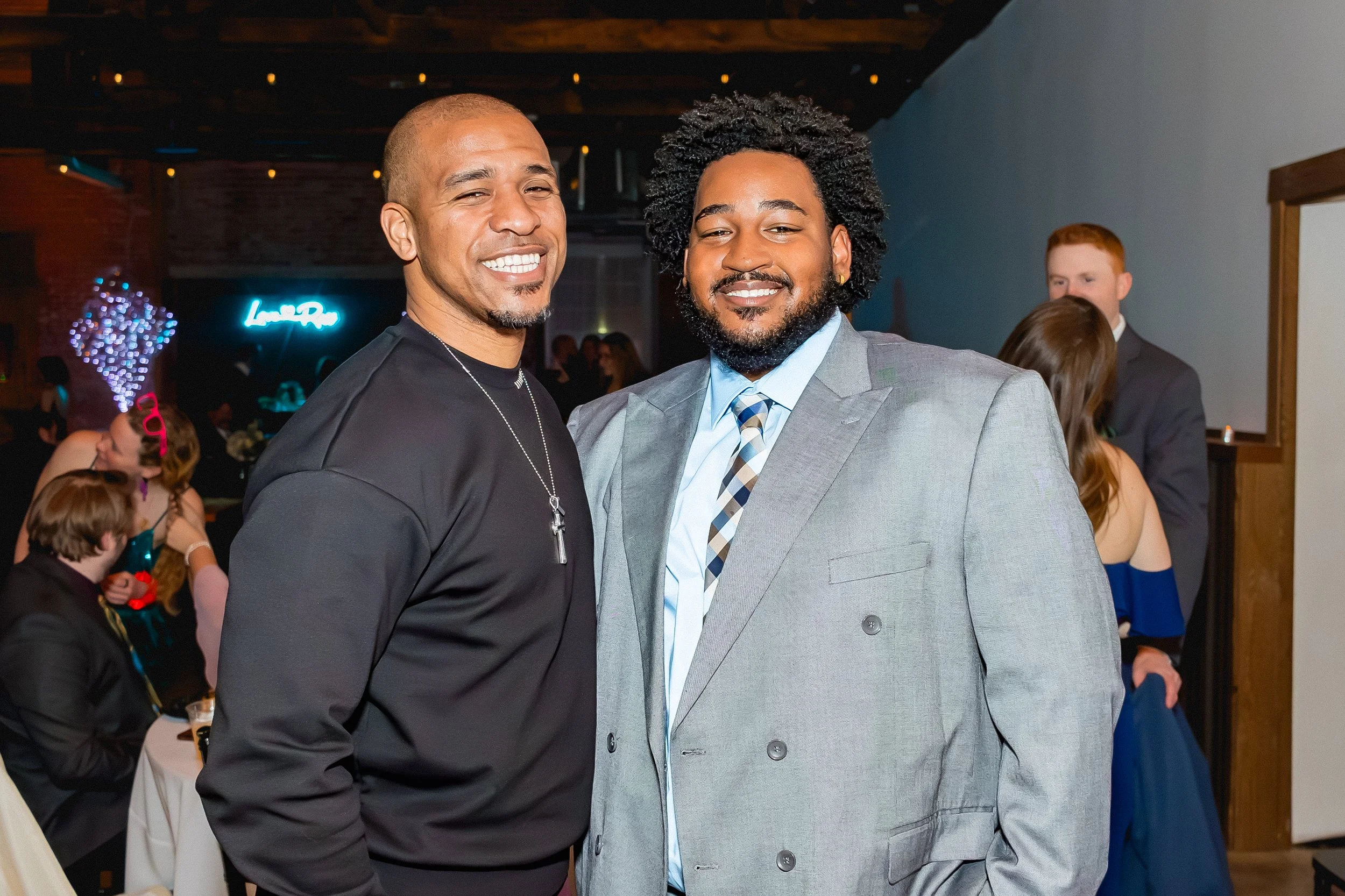 Two men smiling at an event, with other attendees in the background, one wearing a dark shirt with a necklace and the other in a gray suit with a patterned tie.