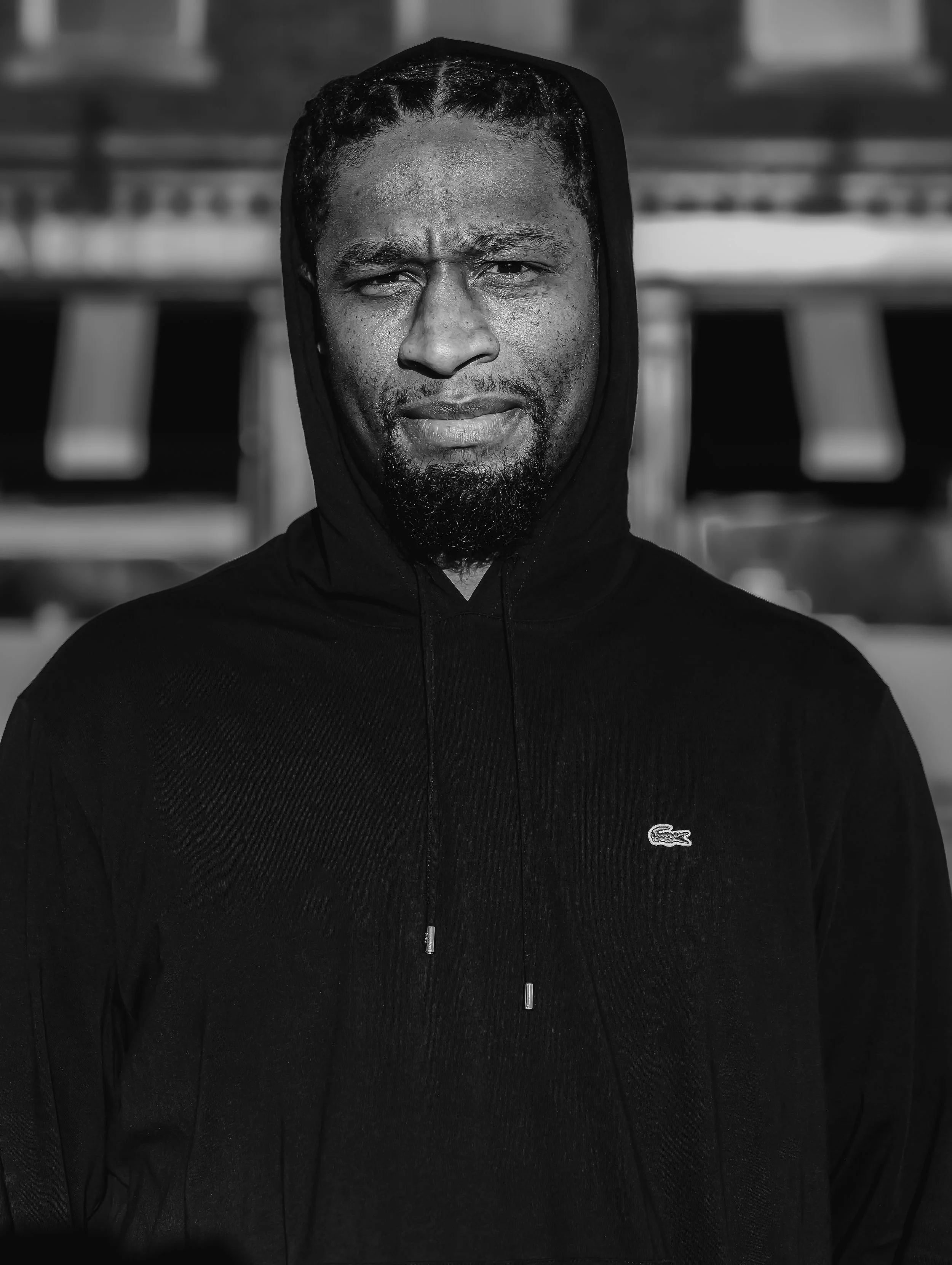 A man wearing a black hoodie with the Lacoste logo, standing outdoors with a blurred background.