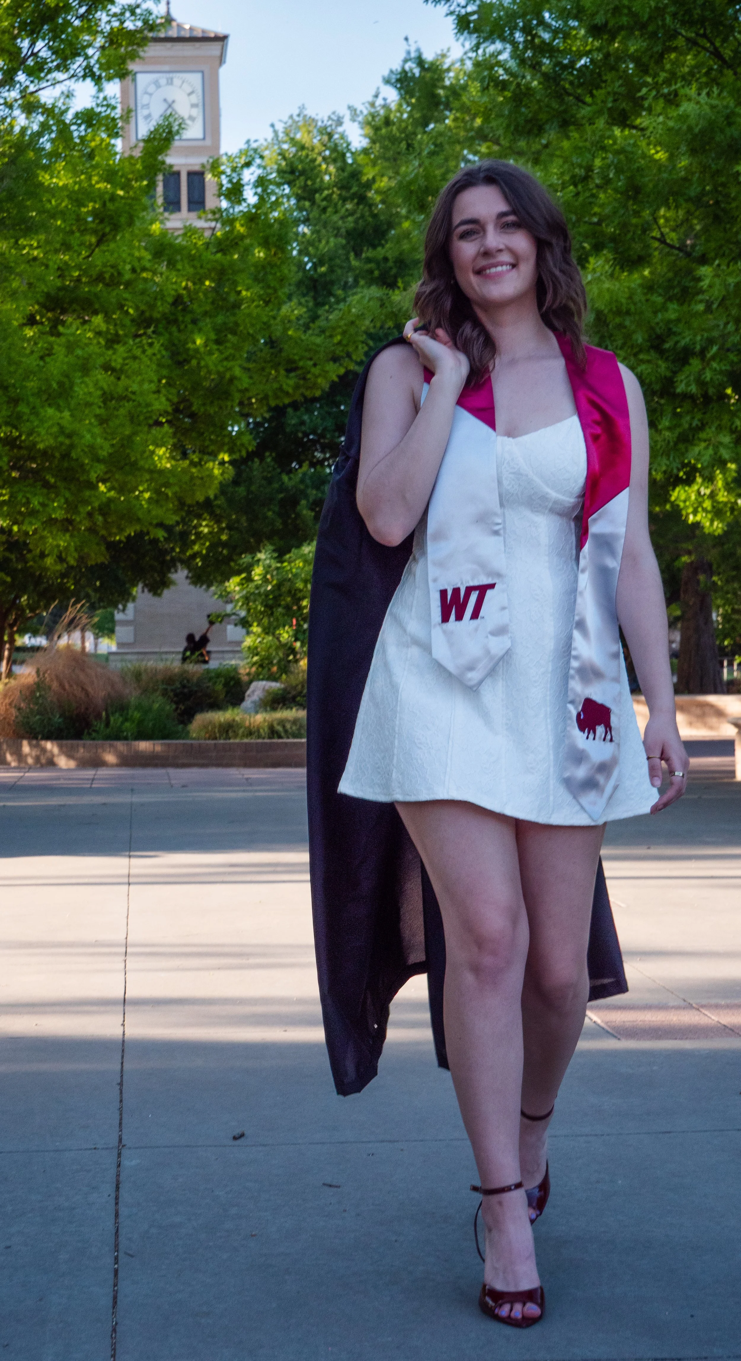 Young woman in a white dress with maroon accents and a graduation stole, walking outdoors in front of green trees, carrying a black gown over her shoulder, smiling, with a clock tower in the background.