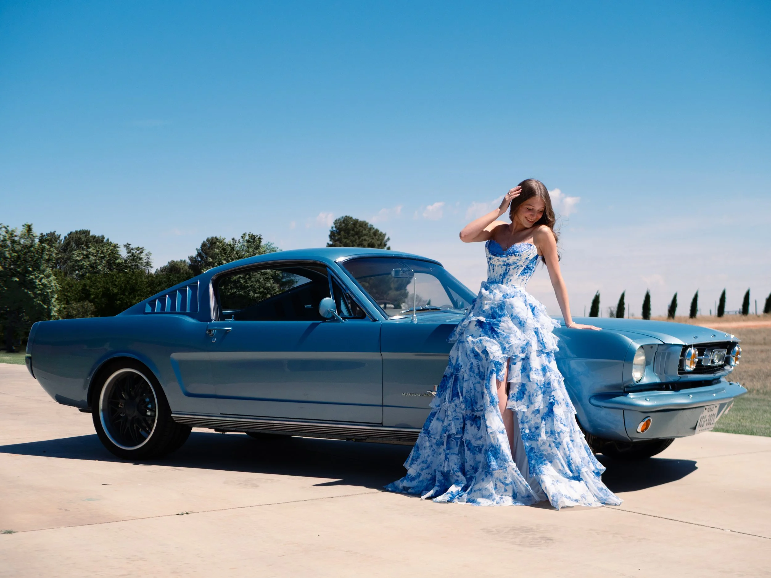 A young woman in a blue and white floral dress standing beside a classic blue Ford Mustang car on a clear day with a blue sky and green trees in the background.