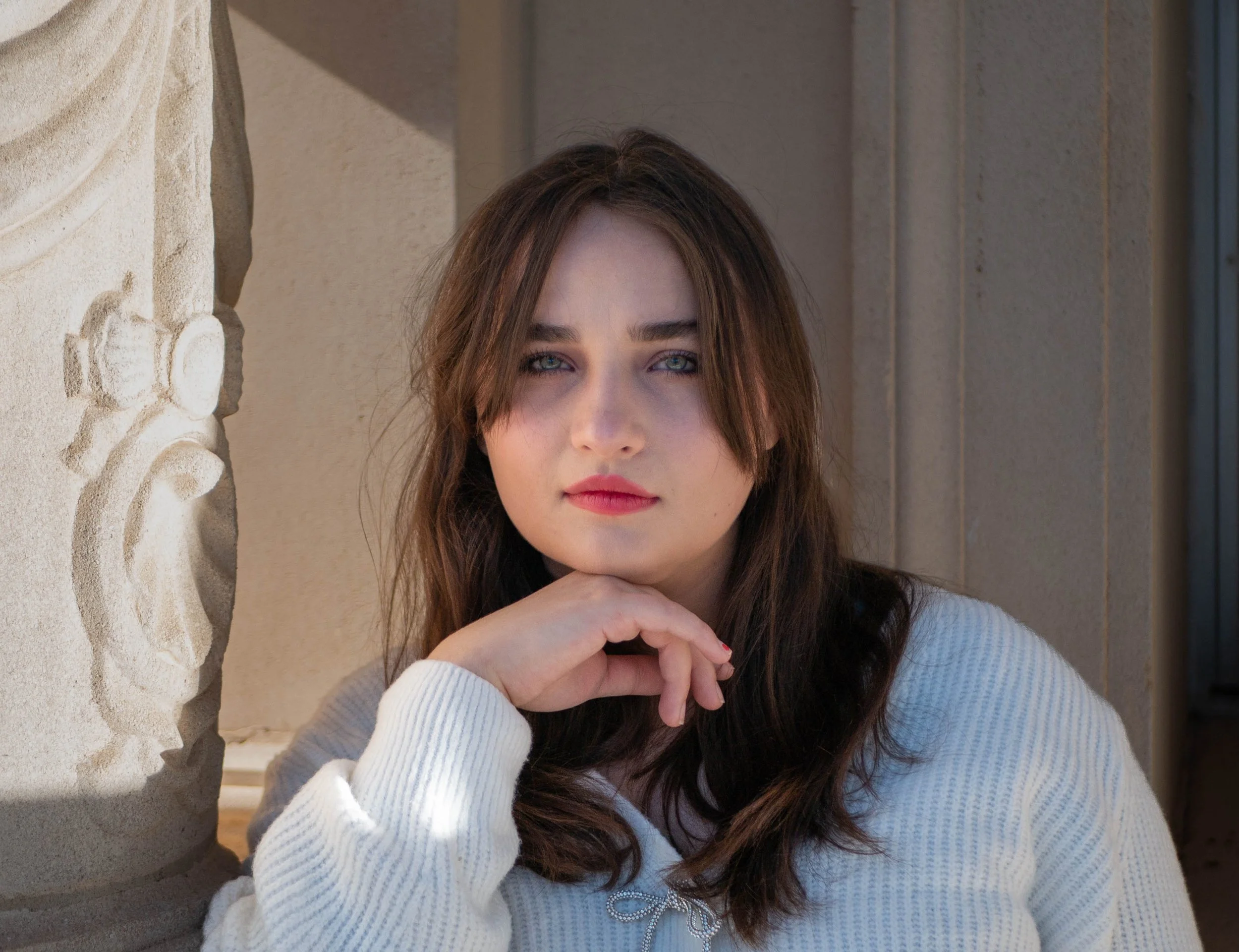 A young woman with long brown hair and blue eyes resting her chin on her hands, wearing a white sweater, standing outdoors near a stone building with a decorative column.