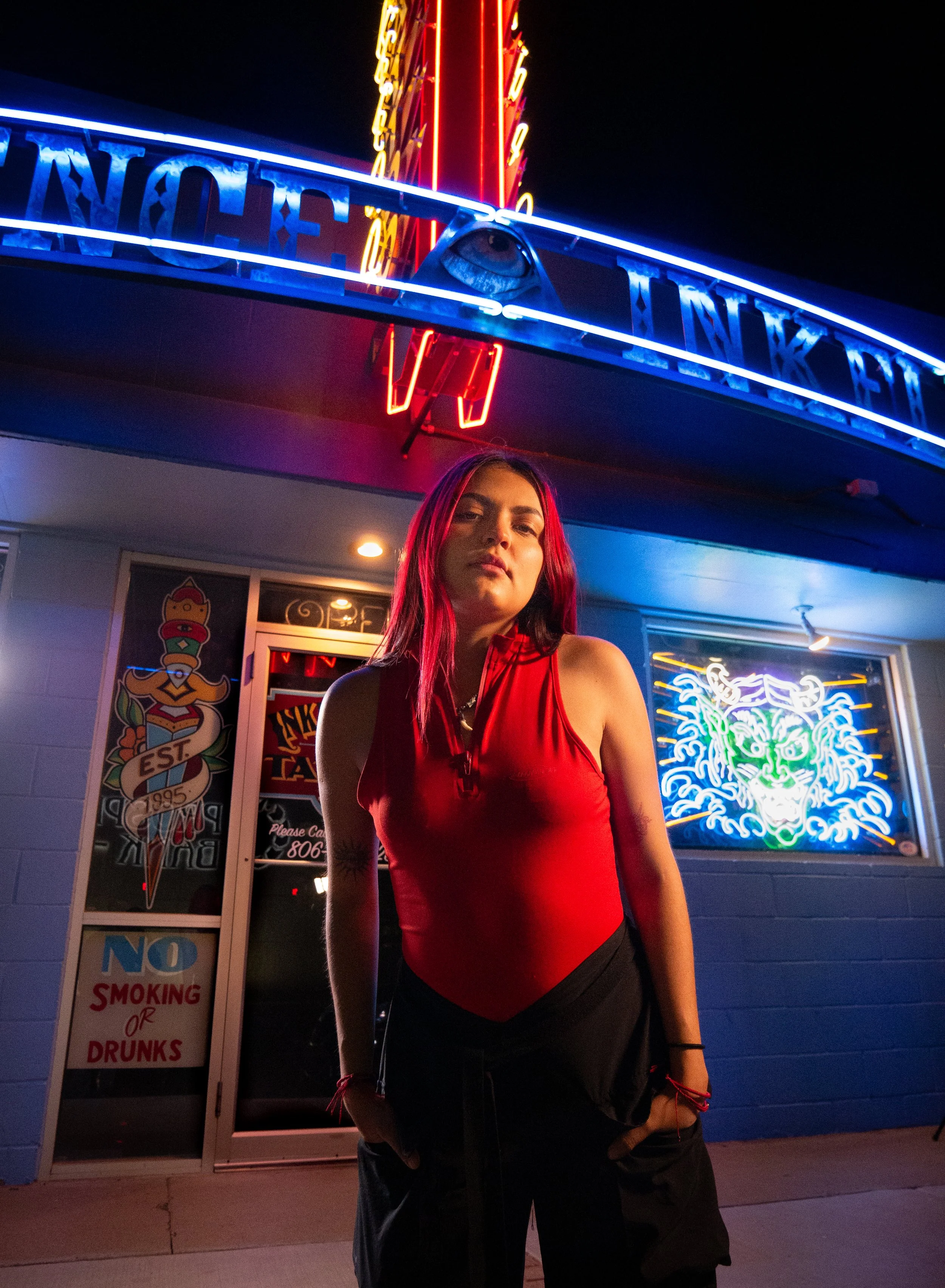 Young woman with red hair wearing a red top and black shorts standing outside at night in front of neon signs, including a dragon's face and a stylized eye, near a door with a 'No Smoking or Drunks' sign.