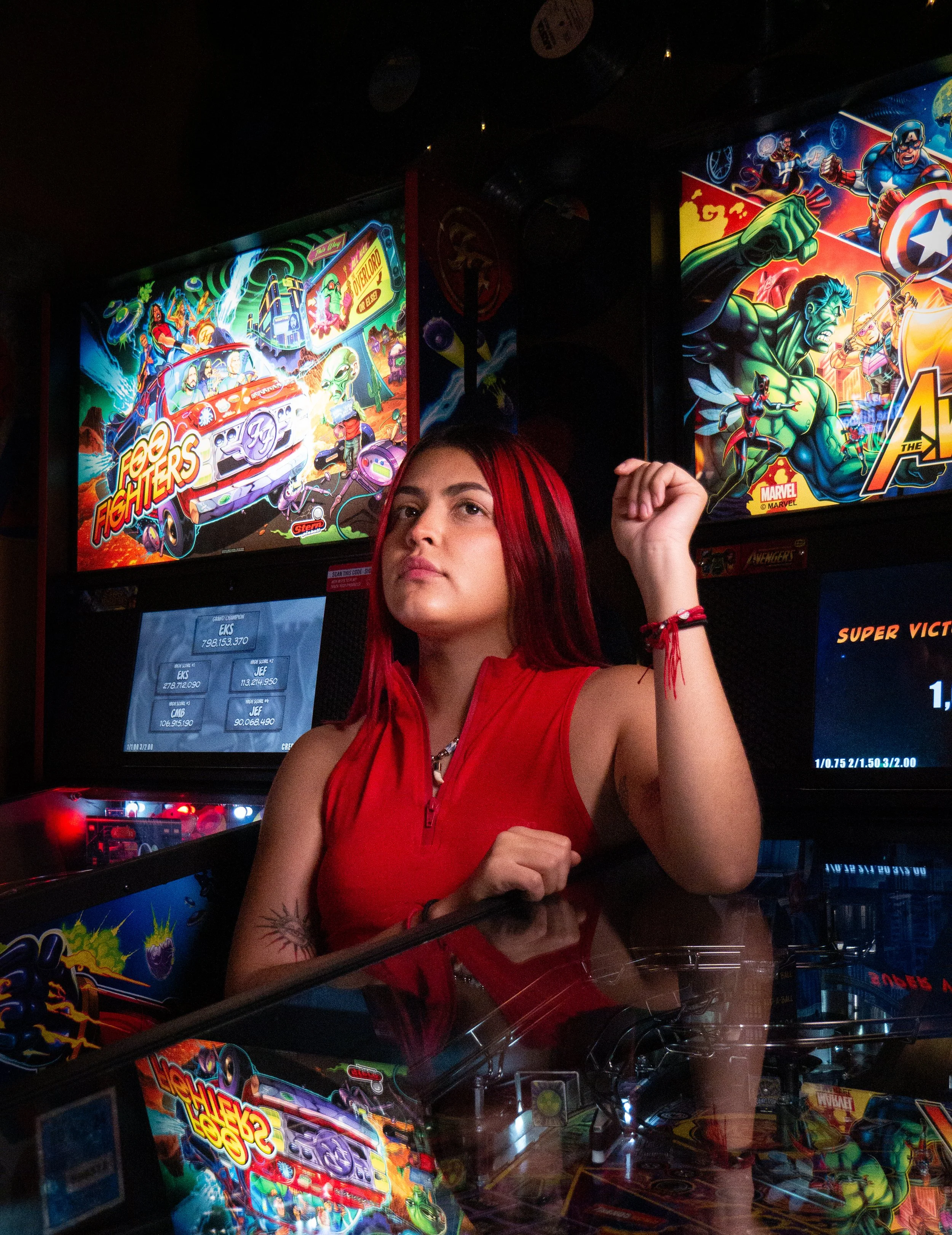 A young woman with red hair and a temporary tattoo on her arm sits in front of arcade pinball machines with colorful Marvel and superhero-themed artwork.