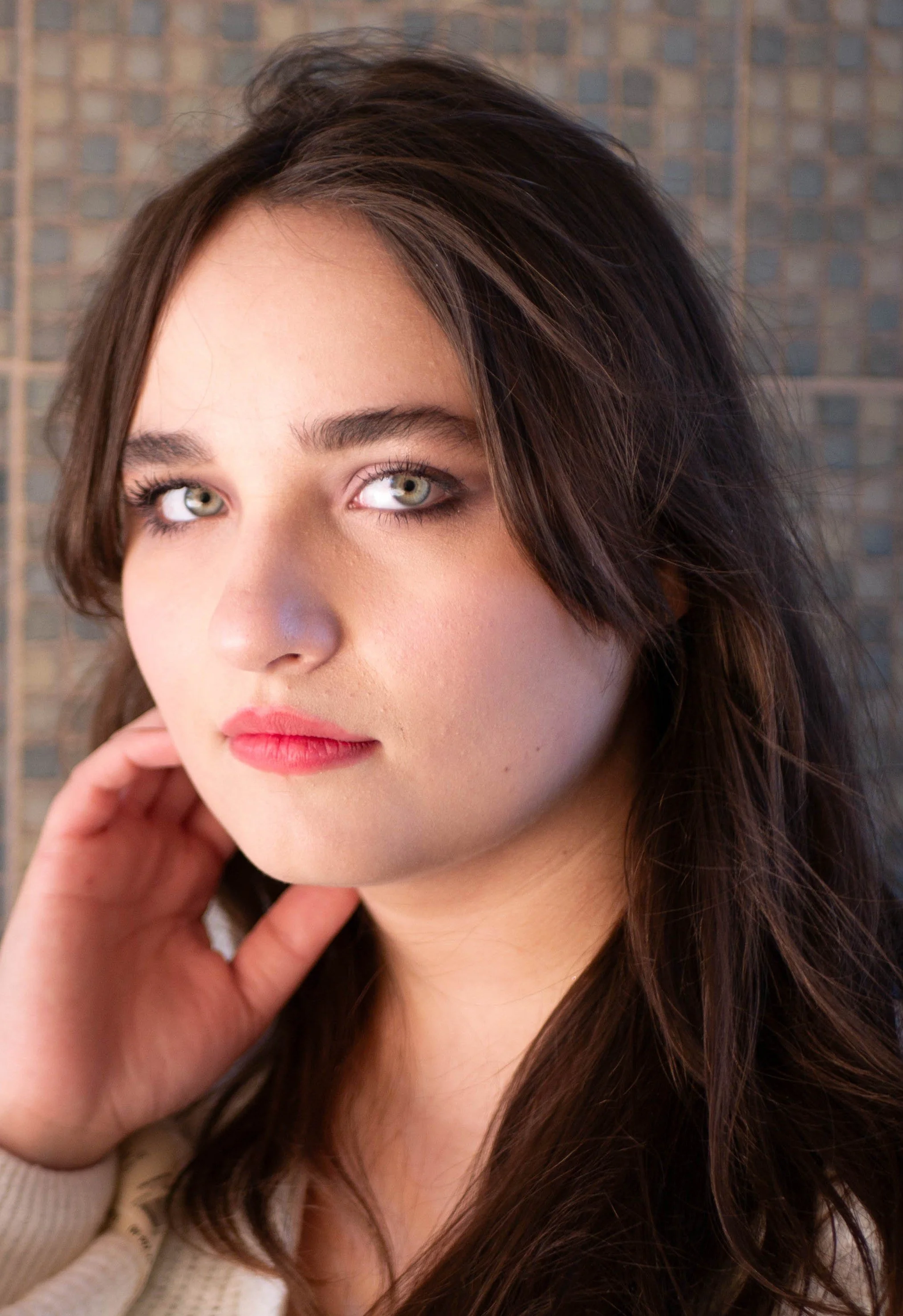 A portrait of a young woman with long wavy brown hair and green eyes, touching her face with one hand, standing in front of a blurred tiled wall.