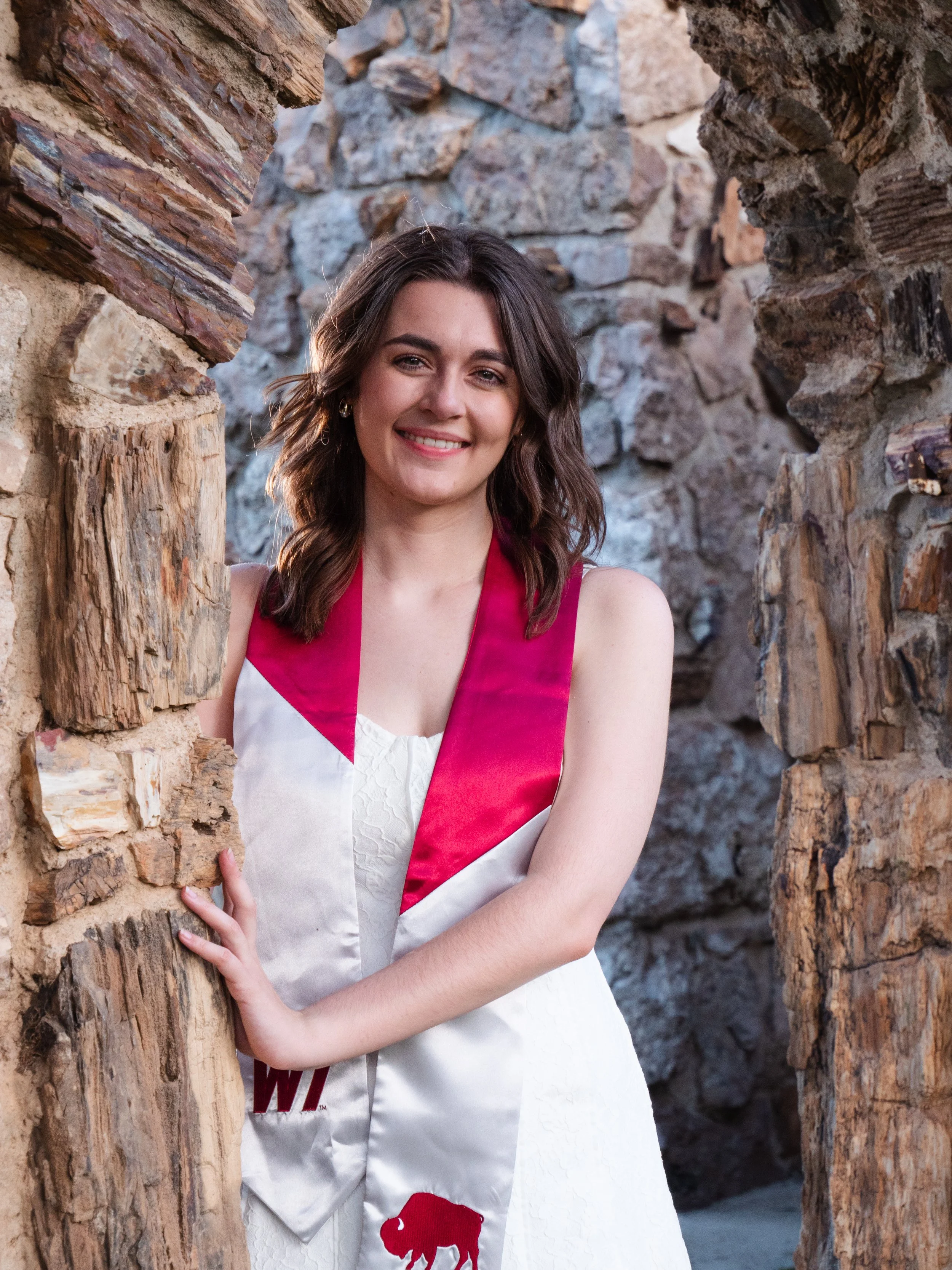 A young woman with shoulder-length brown hair and a smile, wearing a white dress with red accents, leaning against a rustic stone wall.