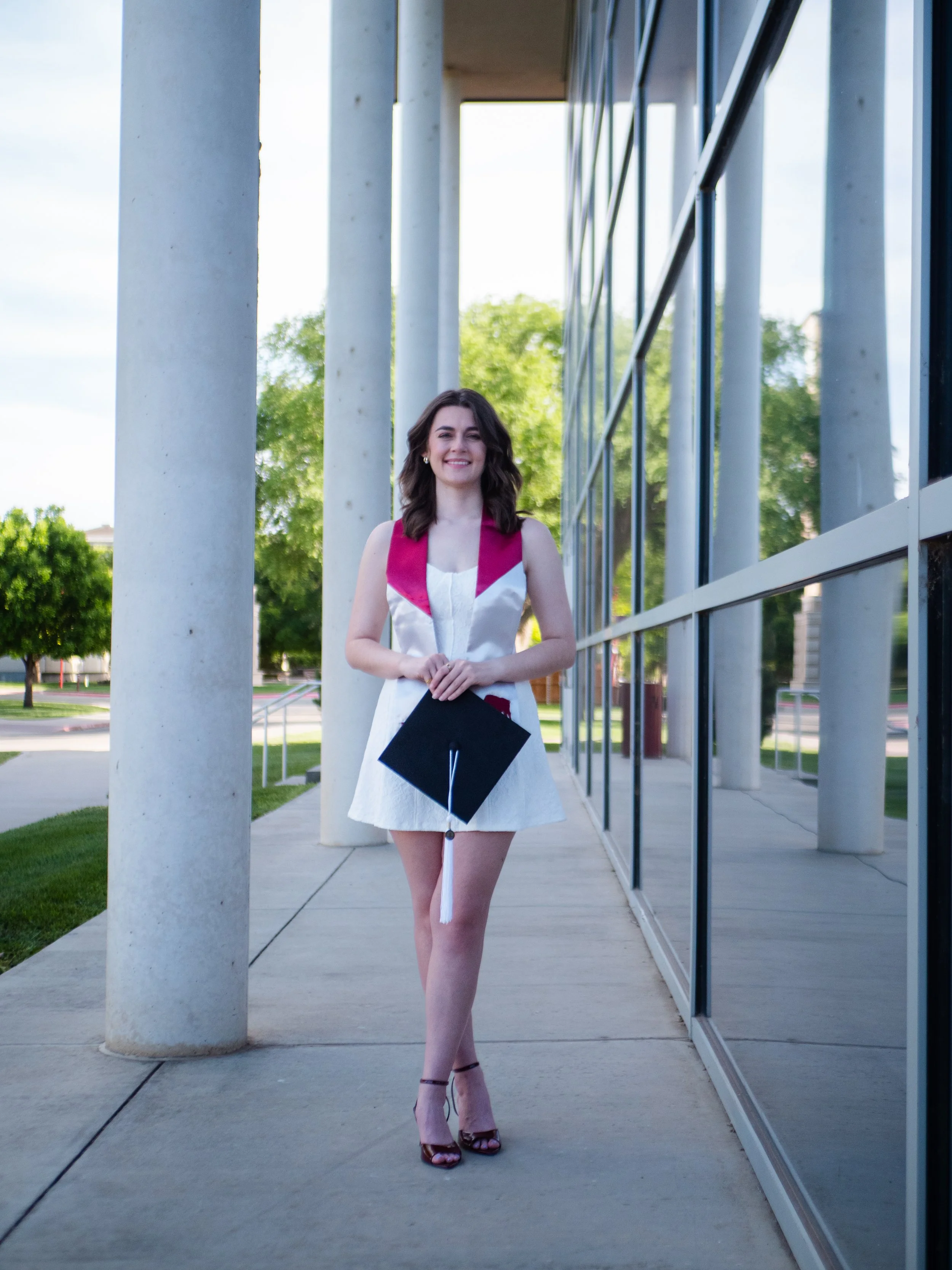 A young woman in a white dress with pink accents and a graduation cap standing outside on a sidewalk with green trees and modern building architecture in the background.
