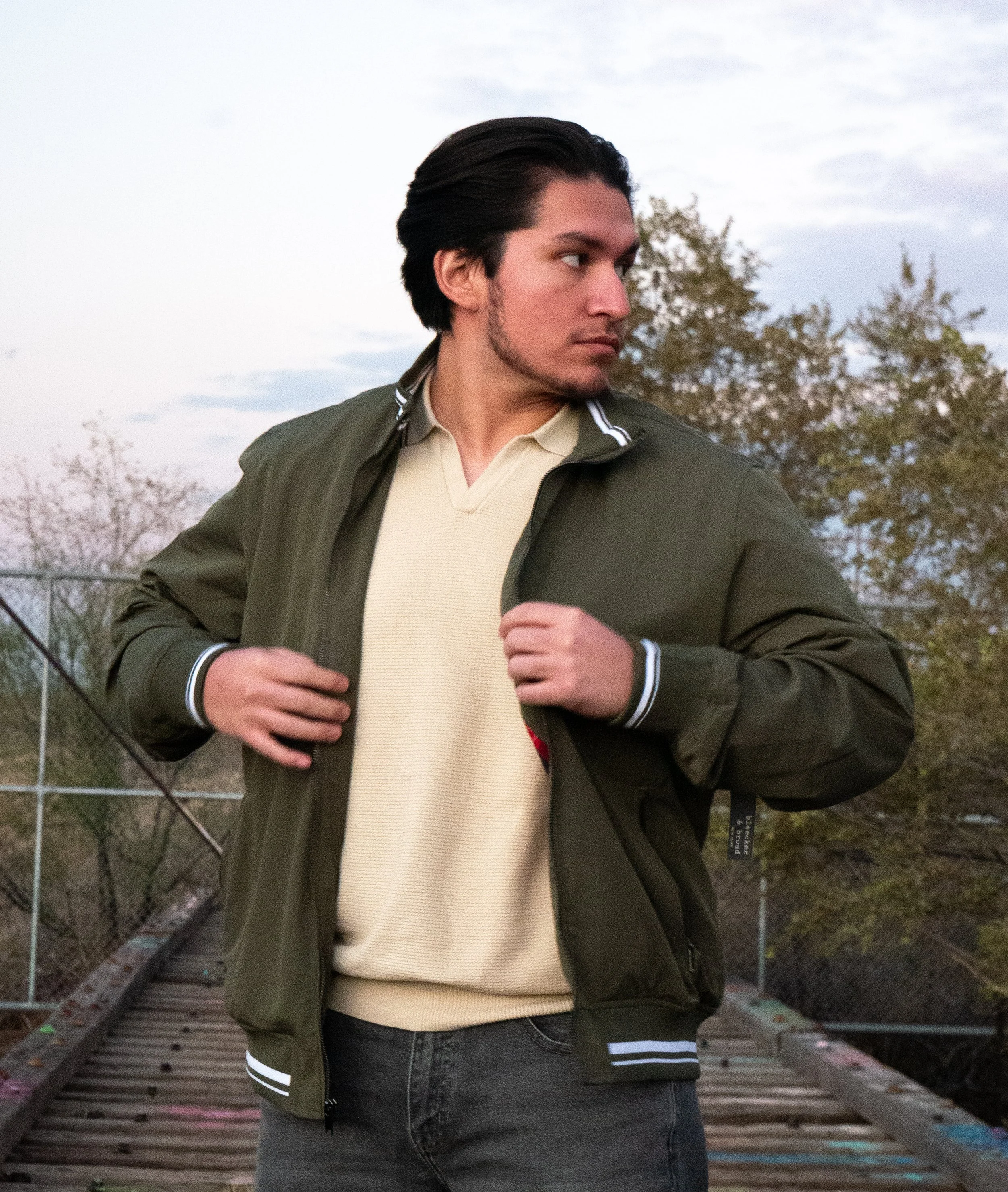 A young man with dark hair, a light beard, wearing a beige sweater, a green jacket, and dark jeans, is standing outdoors with trees and a cloudy sky in the background, captured in profile.