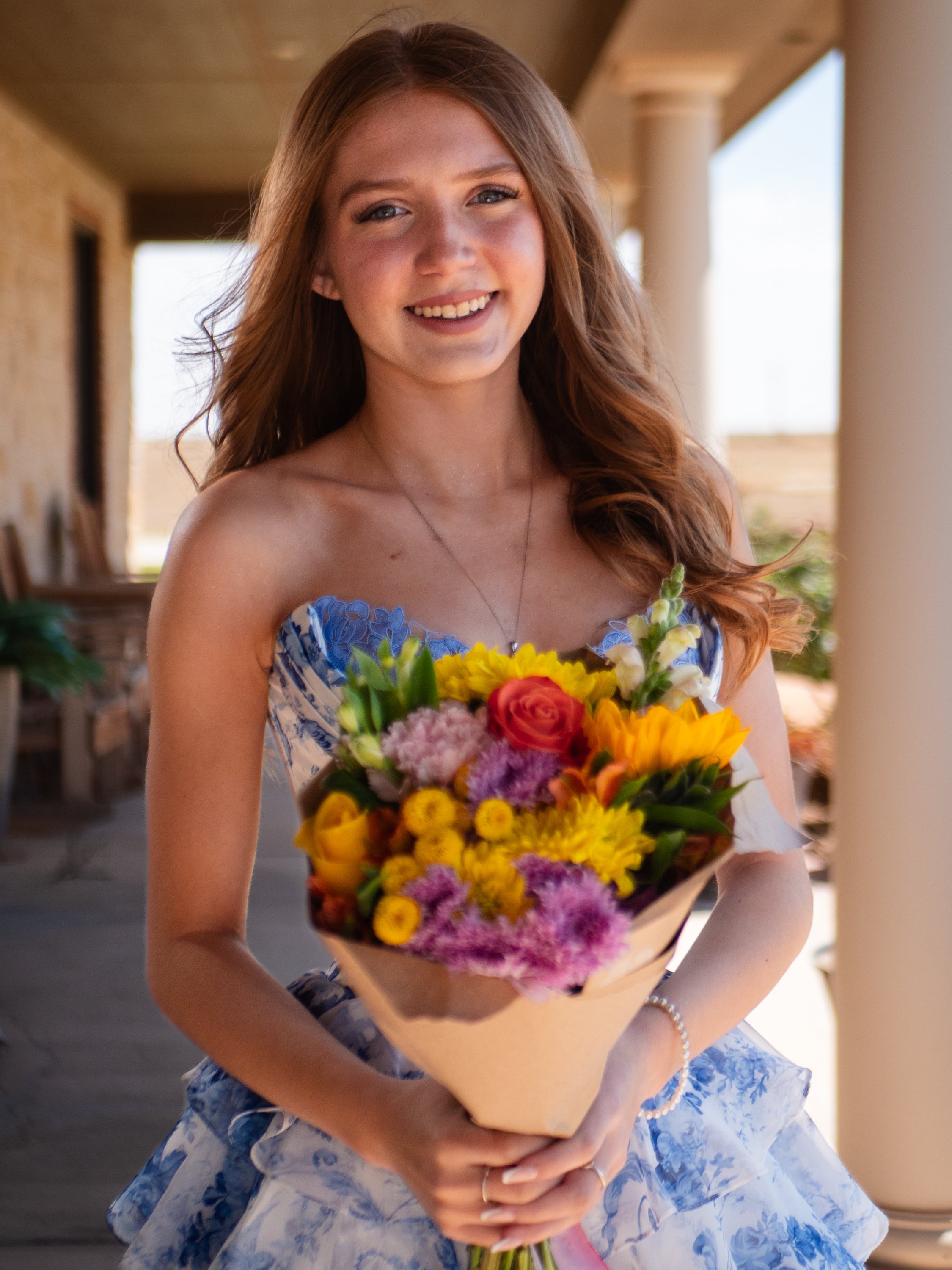 A young woman smiling and holding a colorful bouquet of flowers, wearing a blue and white patterned dress, standing outdoors near a porch with columns.