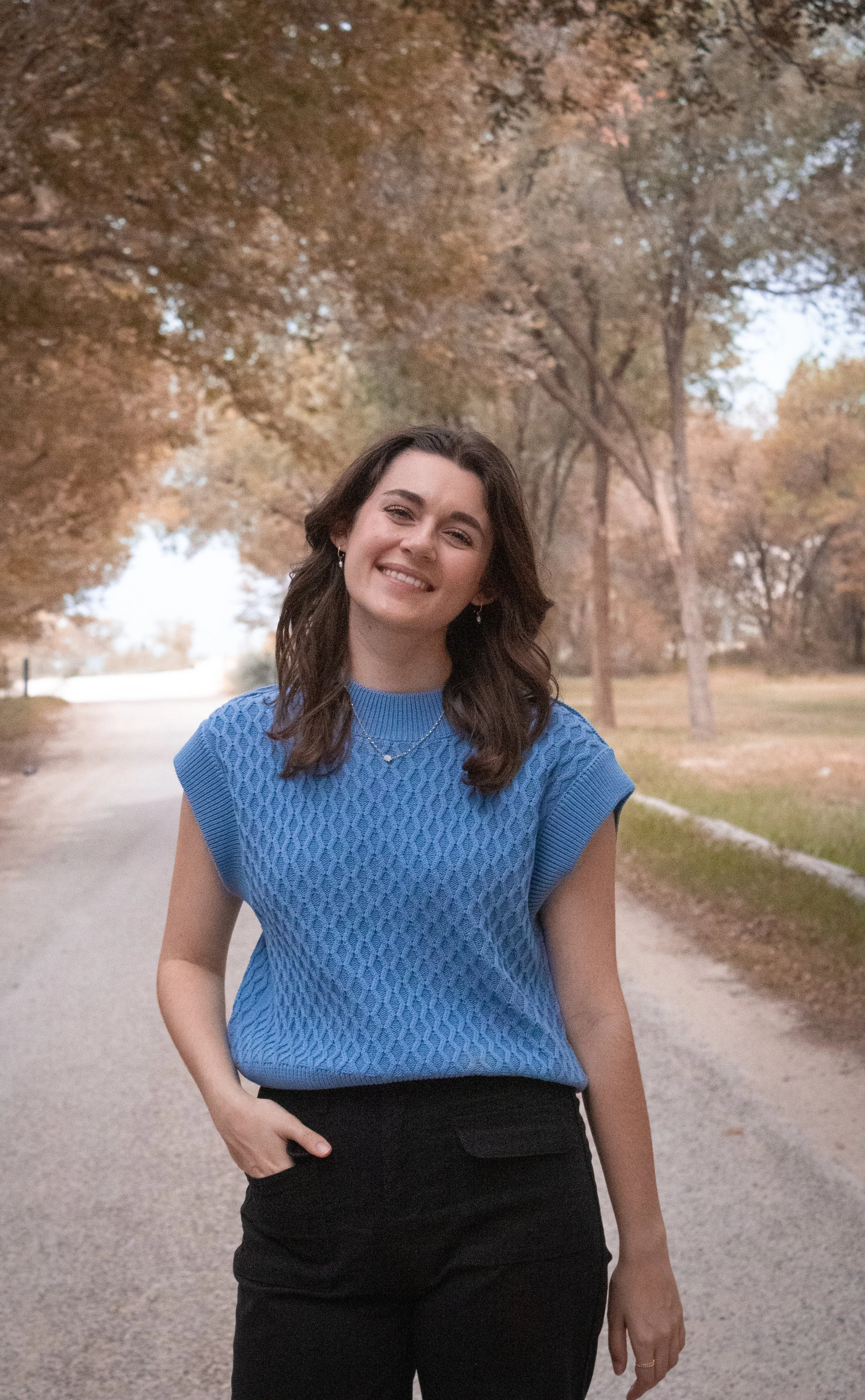 A woman smiling in a park with trees in the background, wearing a blue short-sleeved sweater and black pants.