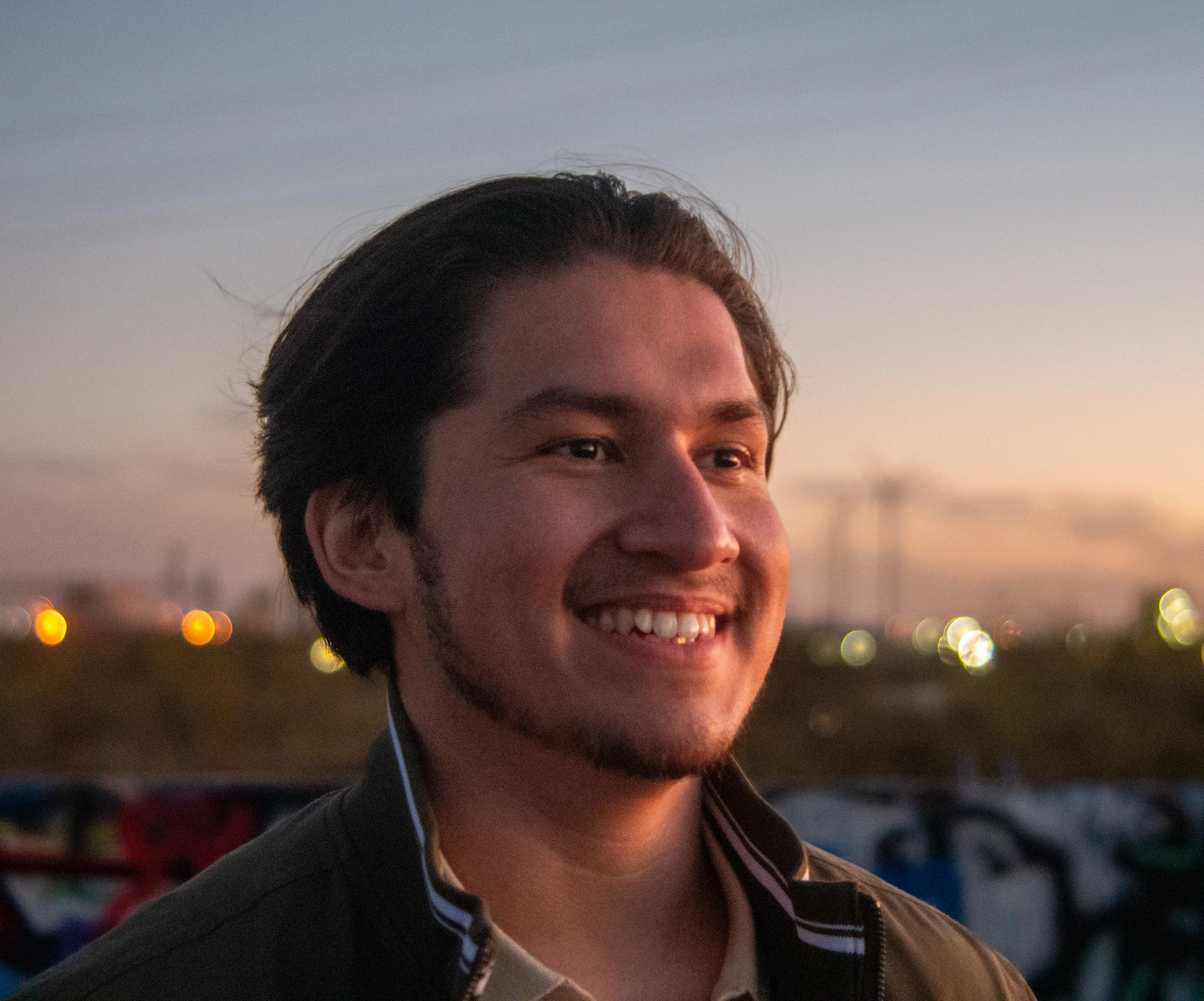 Close-up of a smiling young man with dark hair, wearing earbuds, against a sunset sky with blurred city lights in the background.