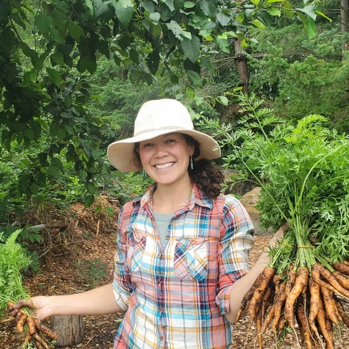 Jasmine Verrall RMT smiling outdoors holding freshly harvested carrots in each hand surrounded by green trees and plants.