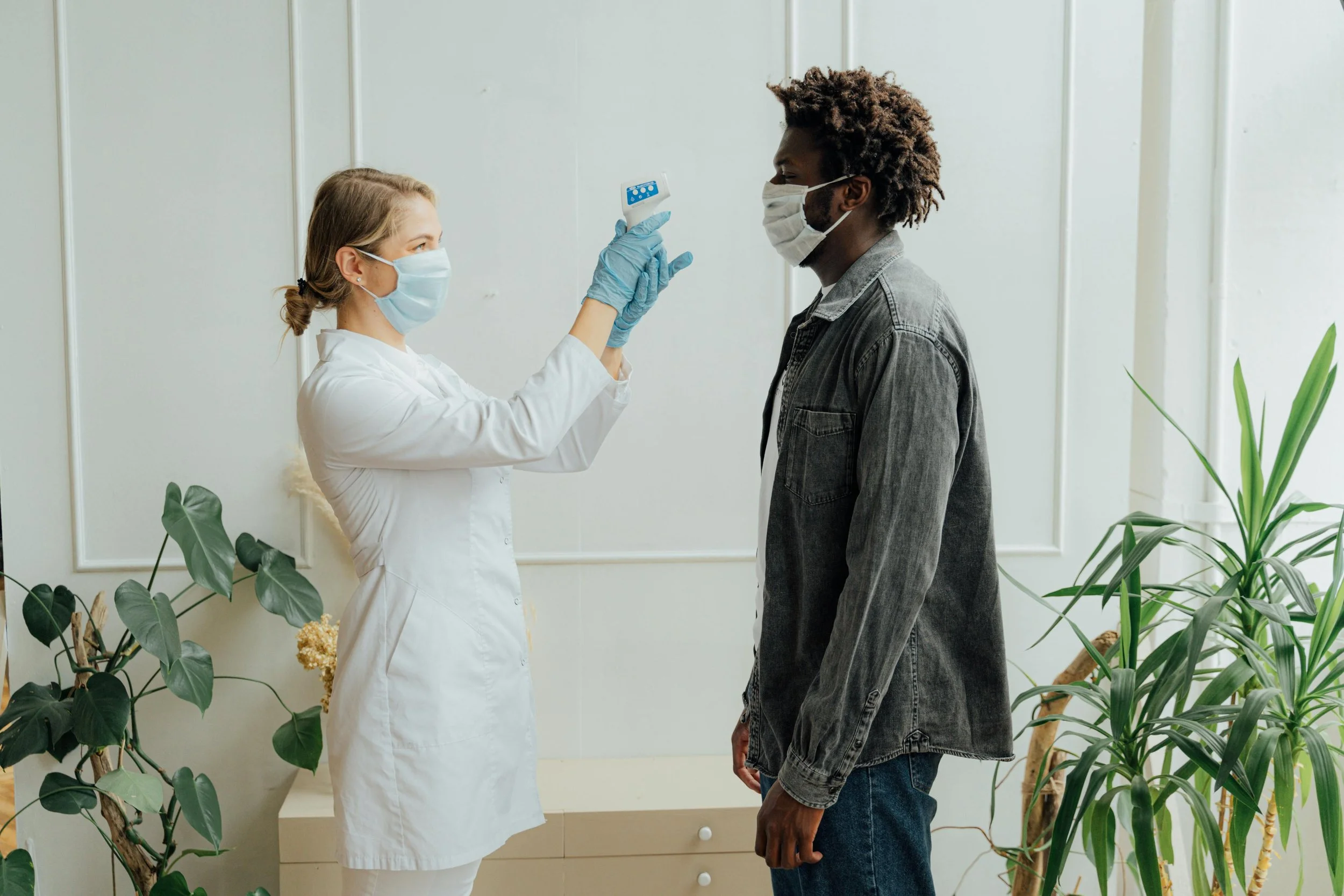 Healthcare professional in a white coat and mask holding a thermometer towards a patient with a mask in a clinical setting with plants visible in the background.