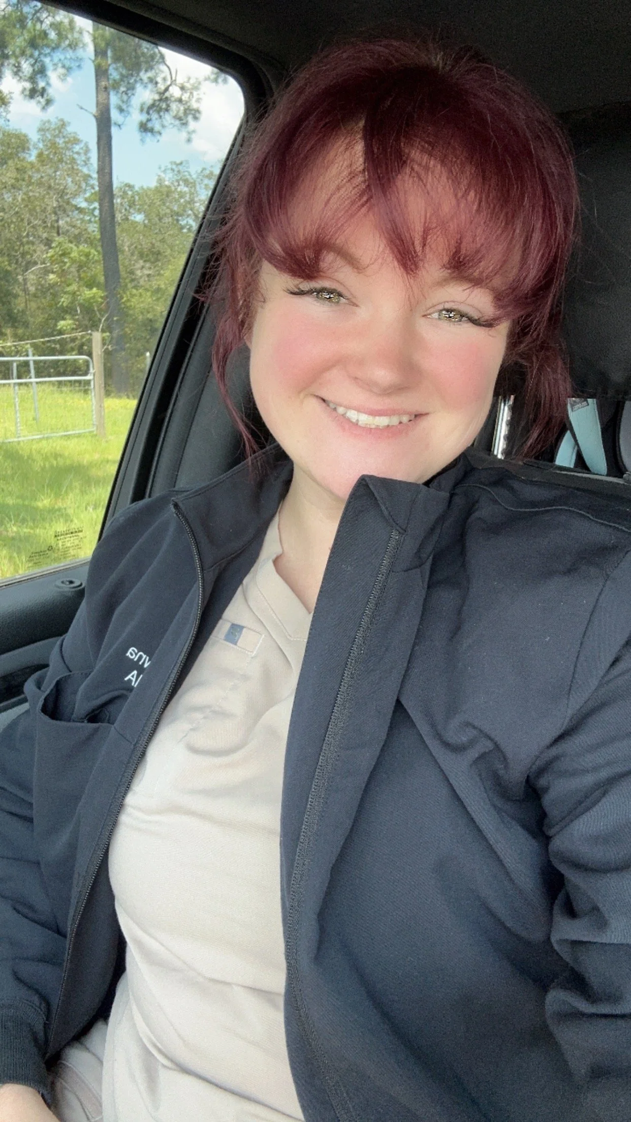 A smiling woman with reddish-brown hair sitting inside a vehicle, wearing a beige uniform and a dark jacket, with greenery and trees outside the window.