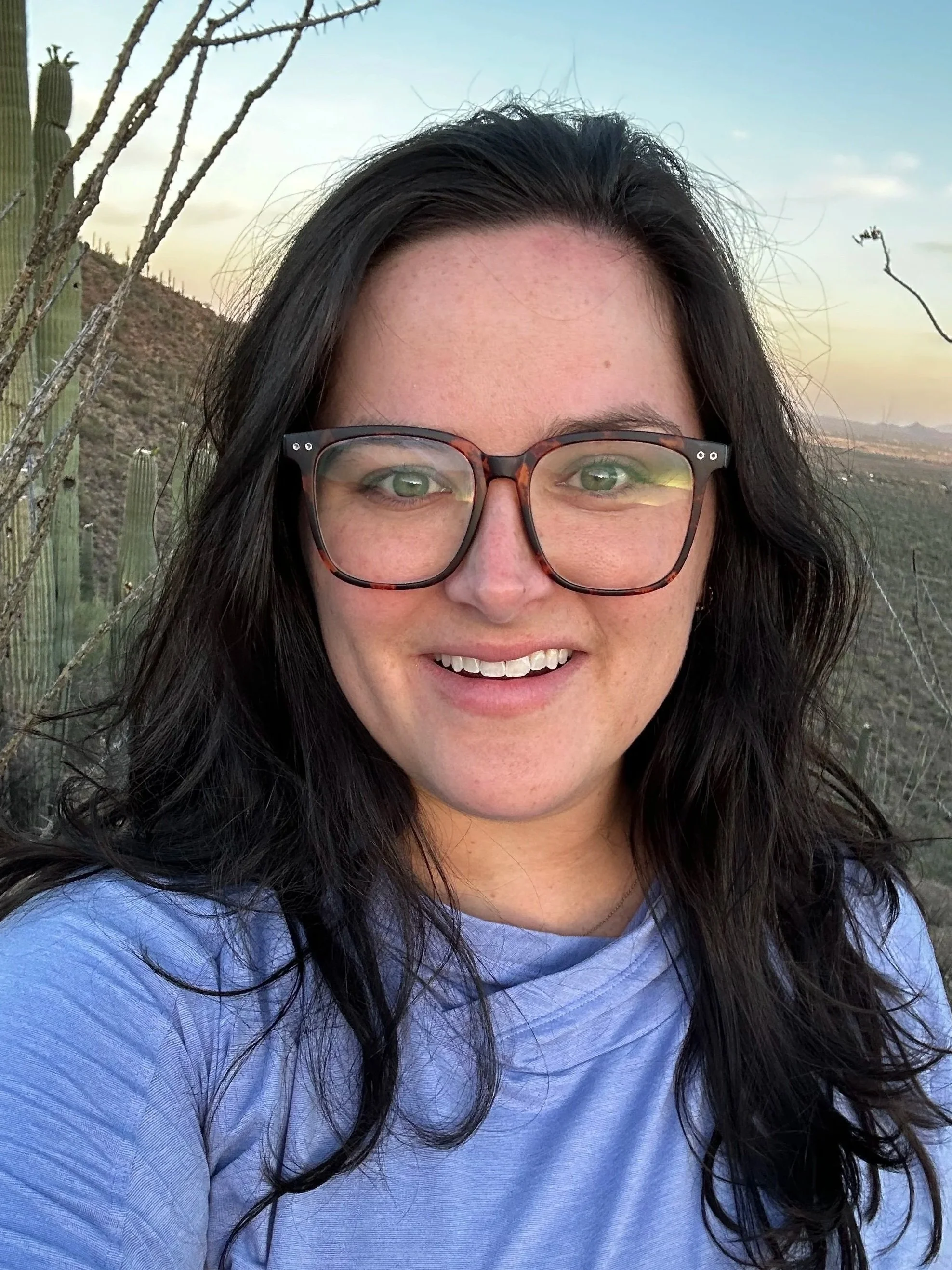 A woman with dark hair and glasses smiling in a desert landscape with cacti and mountains in the background during sunset.