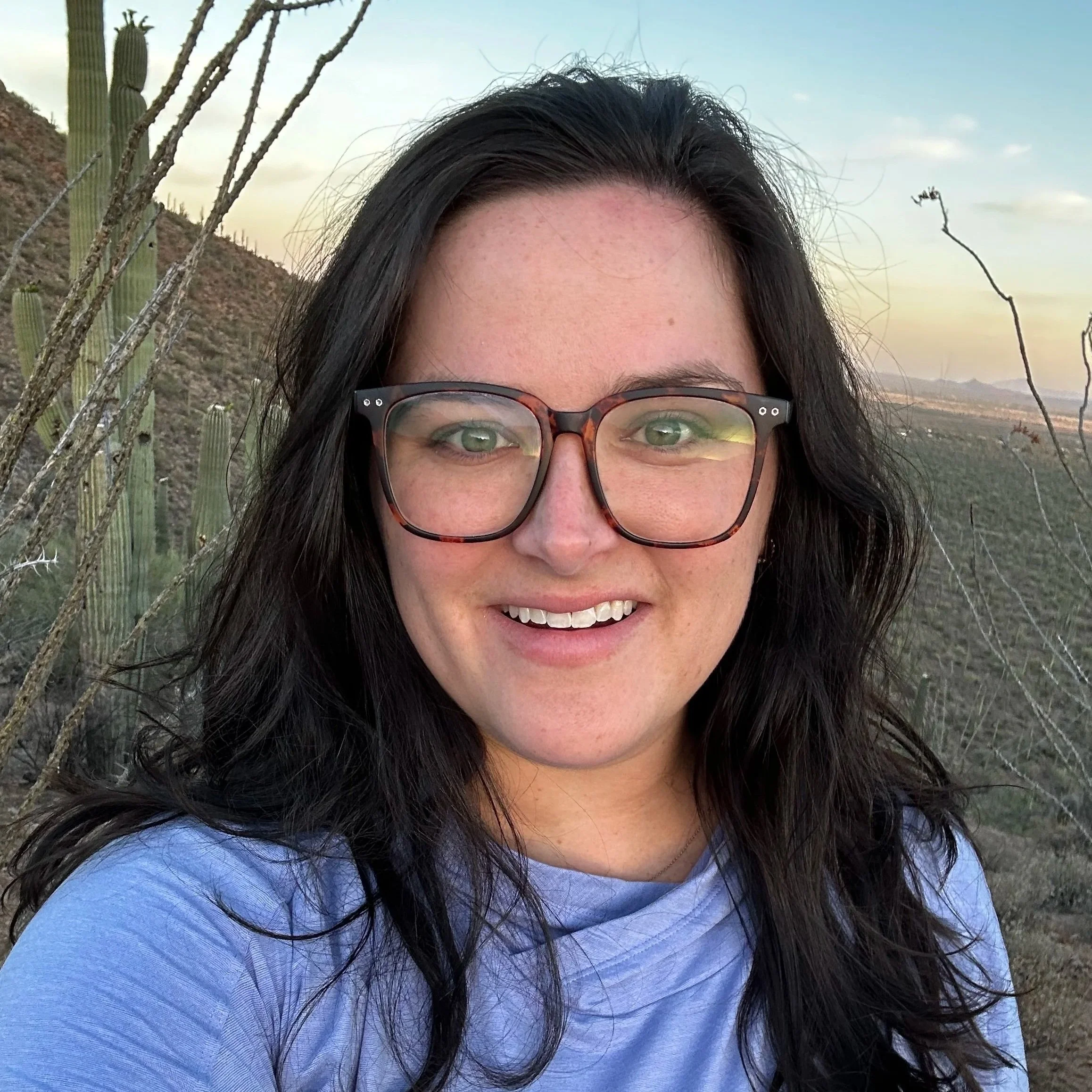 A woman with long dark hair, glasses, and wearing a light purple shirt smiling outdoors in a desert landscape with cacti and hills during sunset.