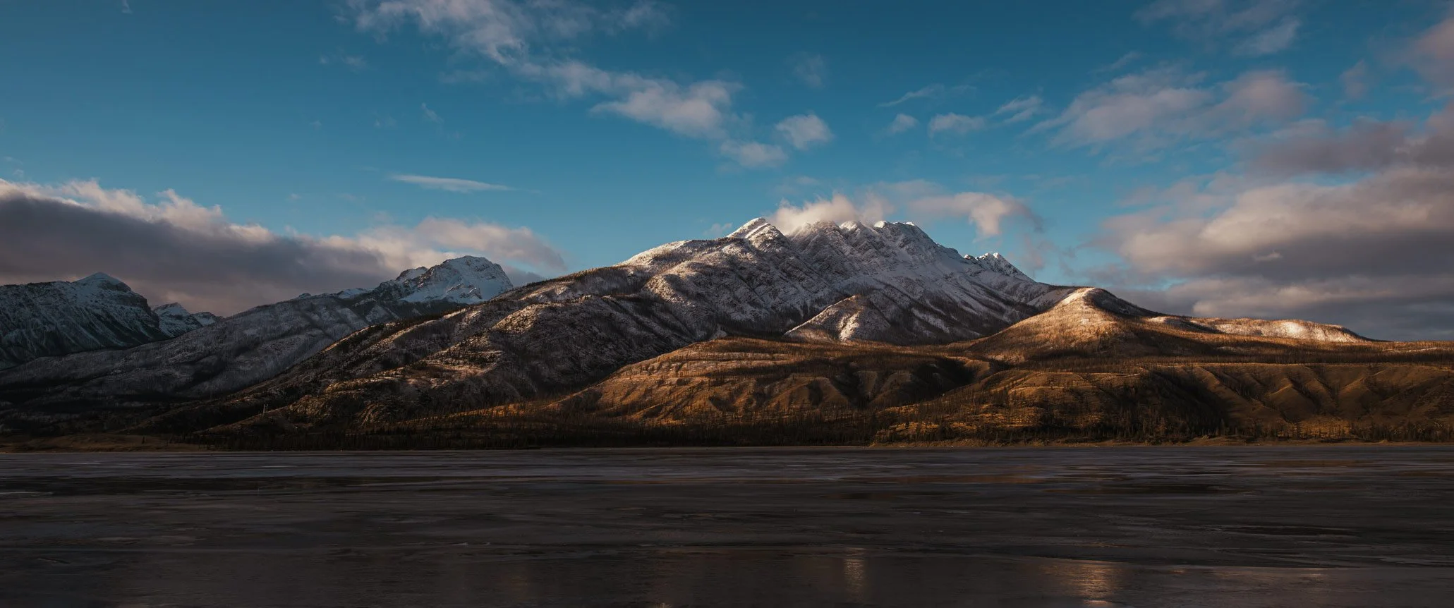 Snow-capped mountains under a partly cloudy sky with a flat dark landscape in the foreground.