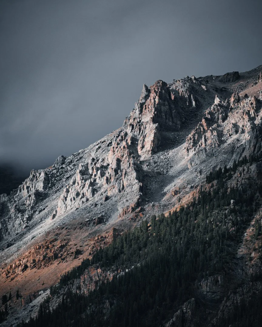 A rugged mountain peak with steep, rocky slopes under a dark, cloudy sky, with a forested area at the base.
