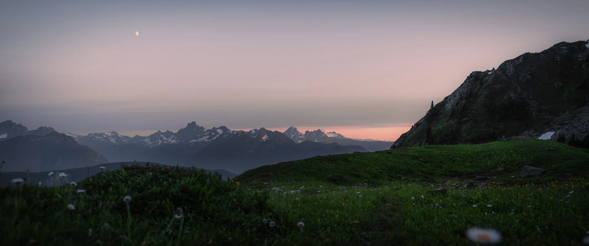 Mountain landscape at dusk with snow-capped peaks, green meadow in foreground, and a visible moon in the sky.