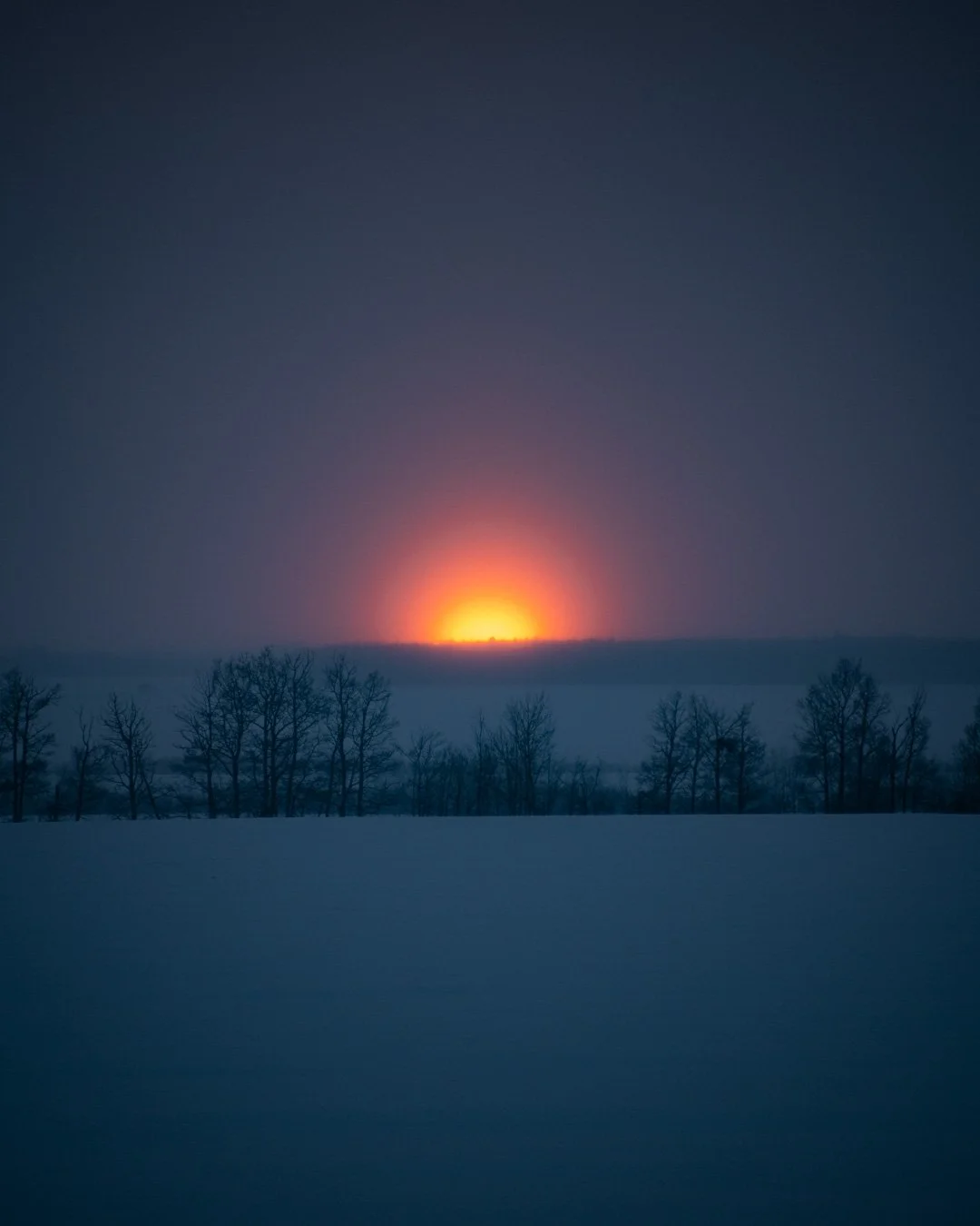 Sunset over a snowy landscape with leafless trees, with orange and purple hues in the sky.