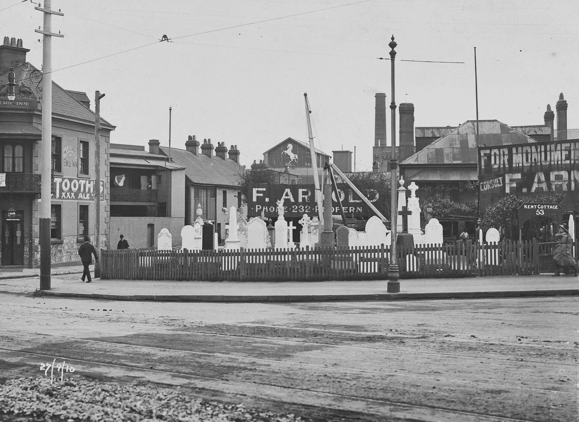 Historical black and white street scene with buildings, power lines, and a small stonemason's yard with white headstones. Several pedestrians walk along the sidewalk.