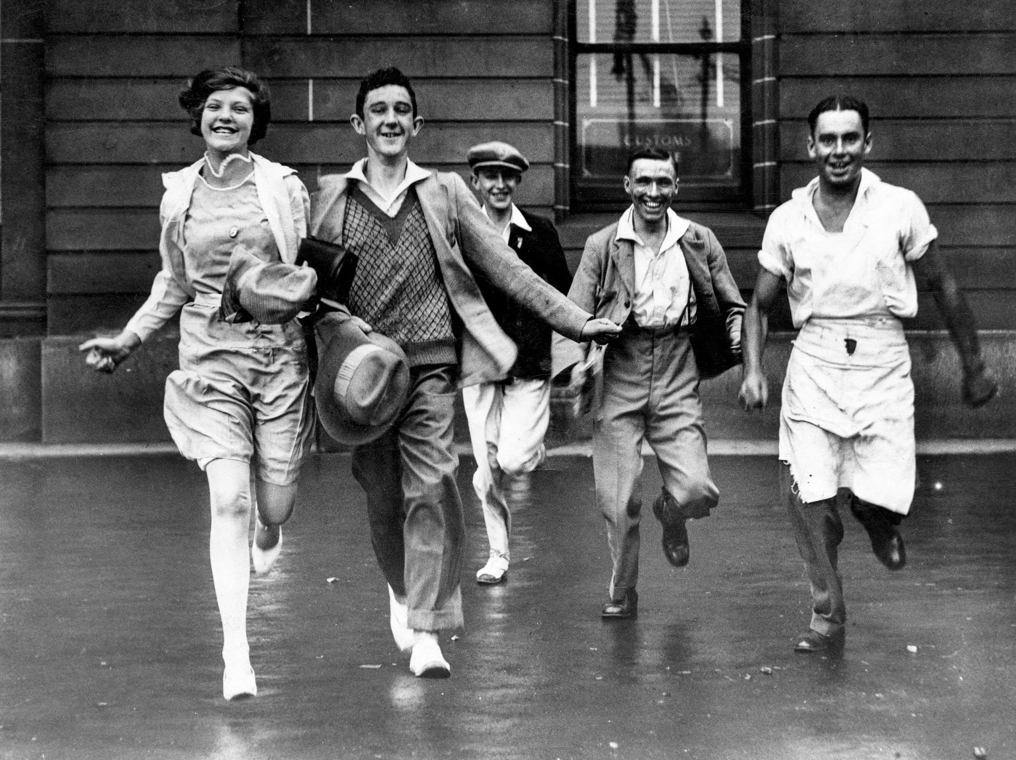 Black and white photo of five people joyfully running and laughing outdoors in front of a wooden building.