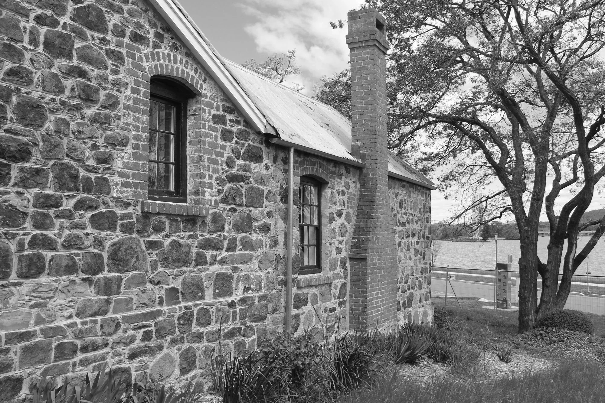 Black and white photo of an old stone house with a chimney, two windows, and a metal roof, with a tree and a river in the background.
