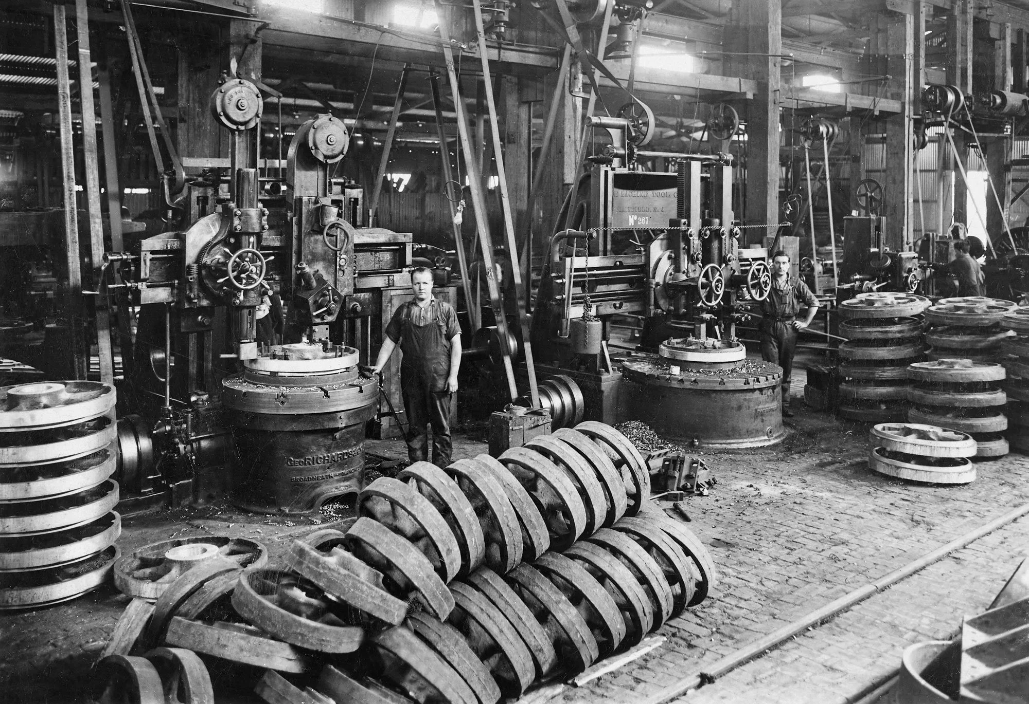 Black and white photograph of workers in a large industrial factory operating heavy machinery and surrounded by stacks of large circular metal parts.