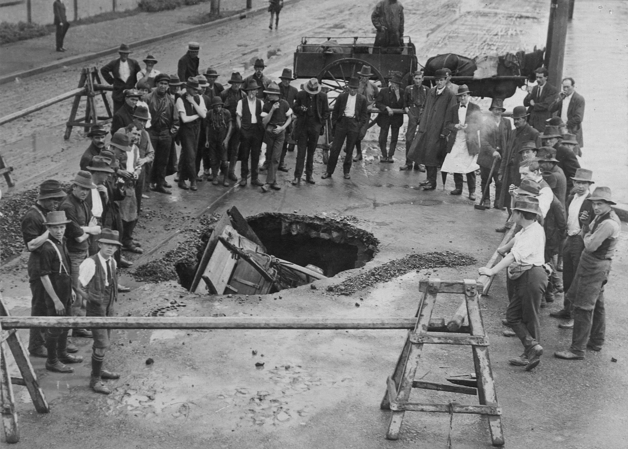 A large group of people, including men and boys, gathered around a large sinkhole in a city street, with some inspecting the damage and others observing or taking photos.