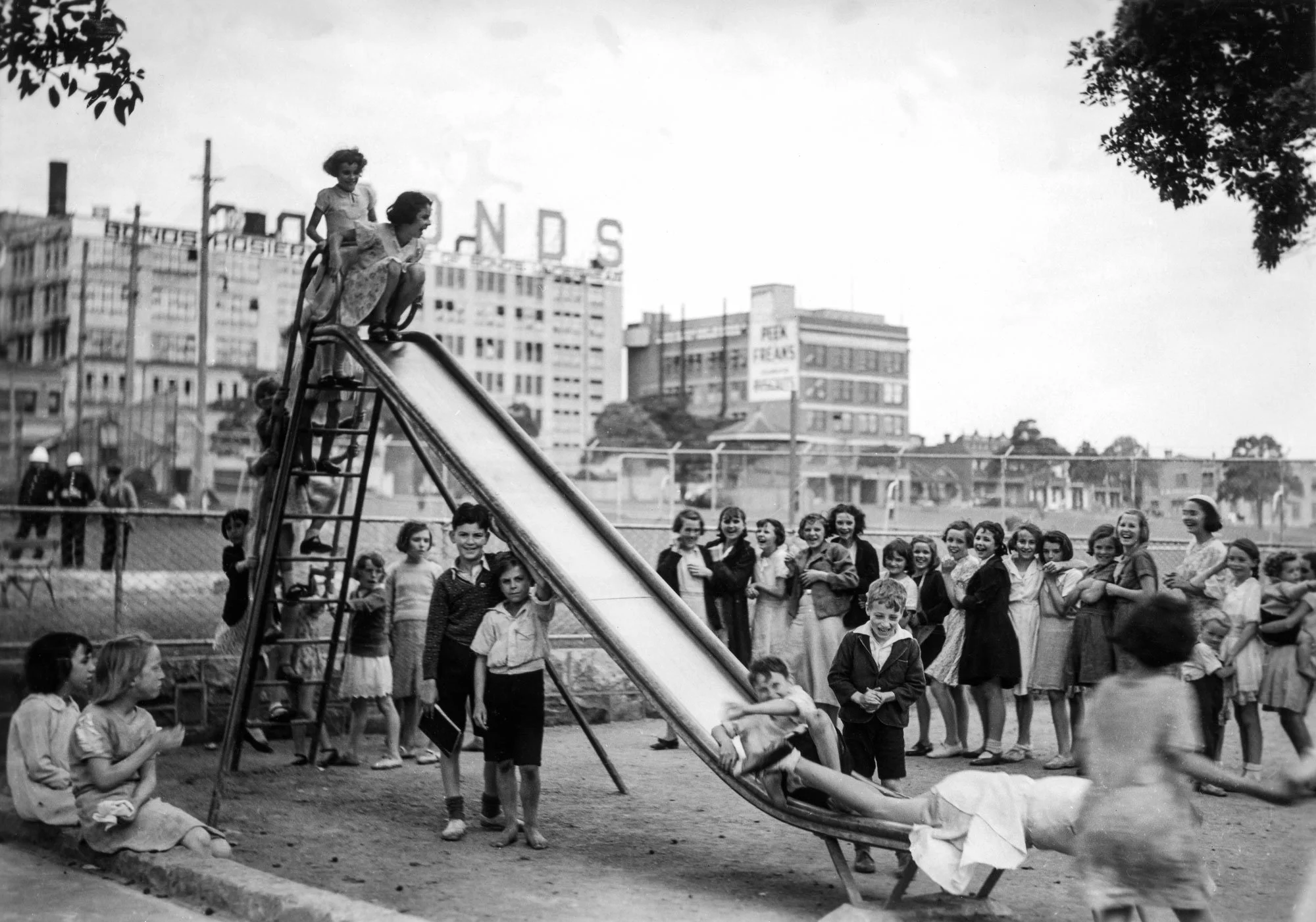 Black and white photograph of children playing on a slide in a park with buildings in the background, some of which have signs and advertising. The children are dressed in clothing typical of the mid-20th century.