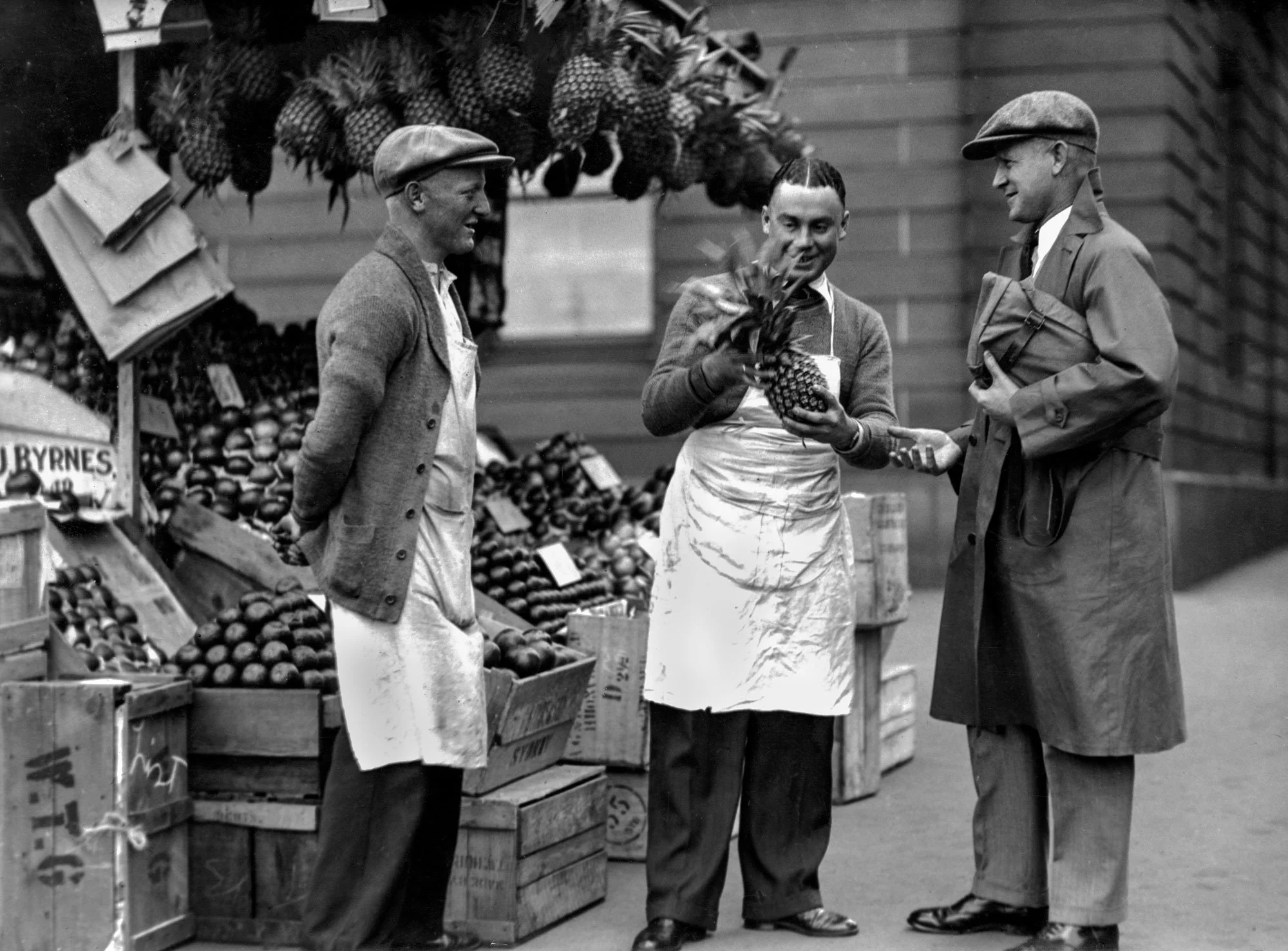 Three people at a fruit stall, with pineapples and other produce in crates behind them. Two men are dressed in coats and hats, one woman is holding a pineapple and smiling, standing between the men.