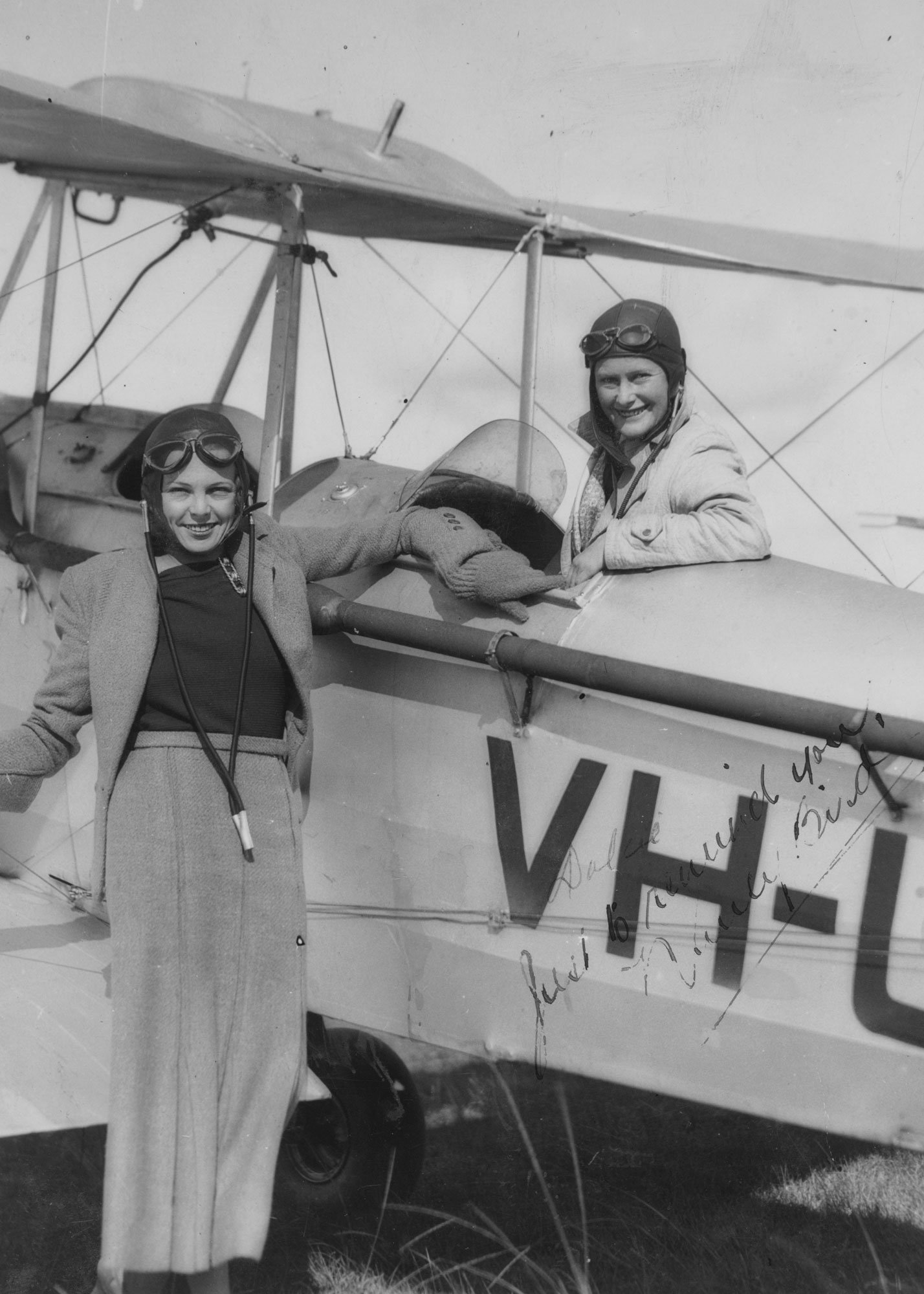 Two women wearing vintage aviator helmets and goggles standing next to a small biplane aircraft, with one woman sitting in the cockpit and the other leaning on the plane.