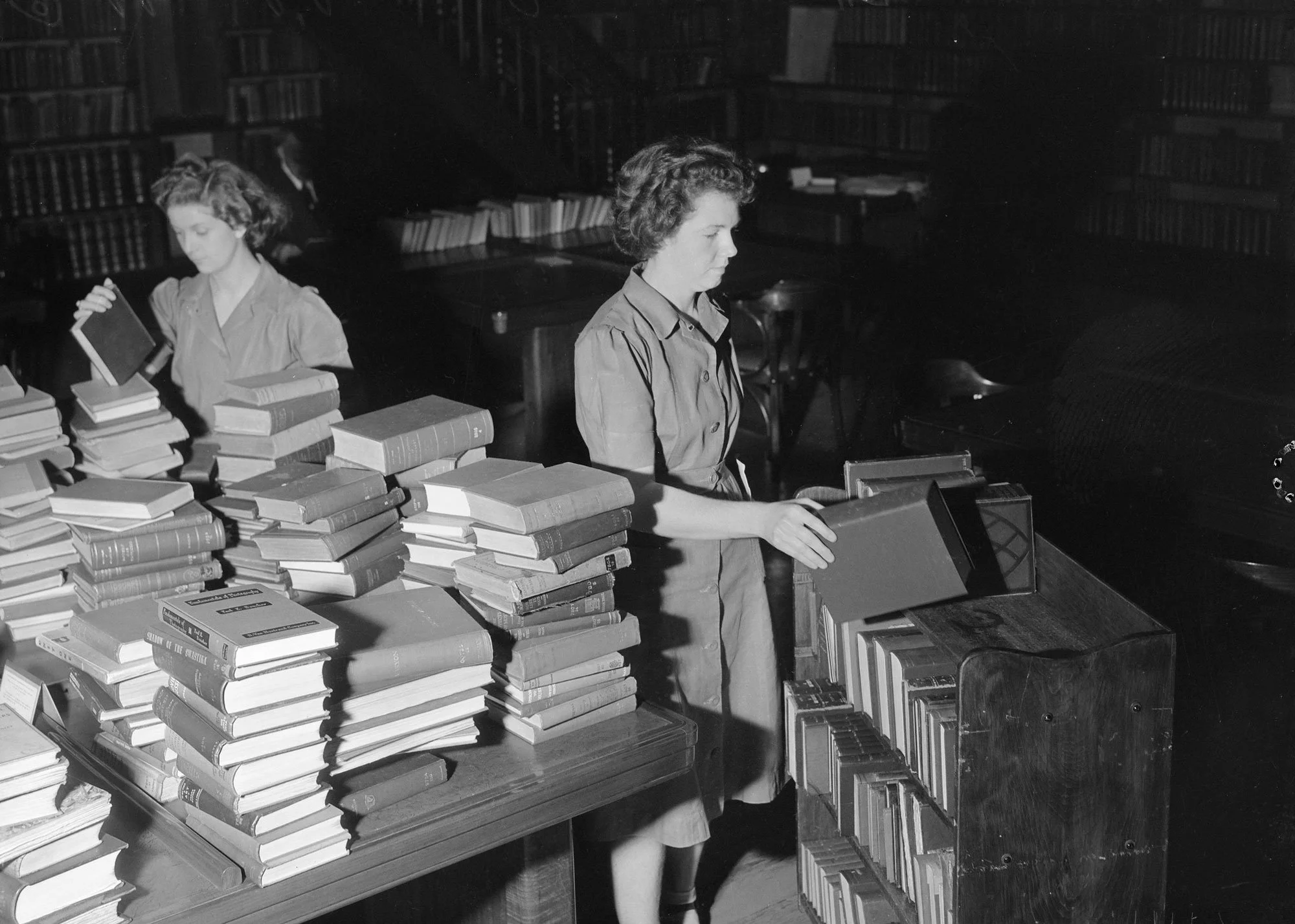 Black and white photo of two women, one in the foreground and the other in the background, organizing books in a library. The woman in the foreground is picking books from a wooden shelf, while the woman in the background is examining a book.