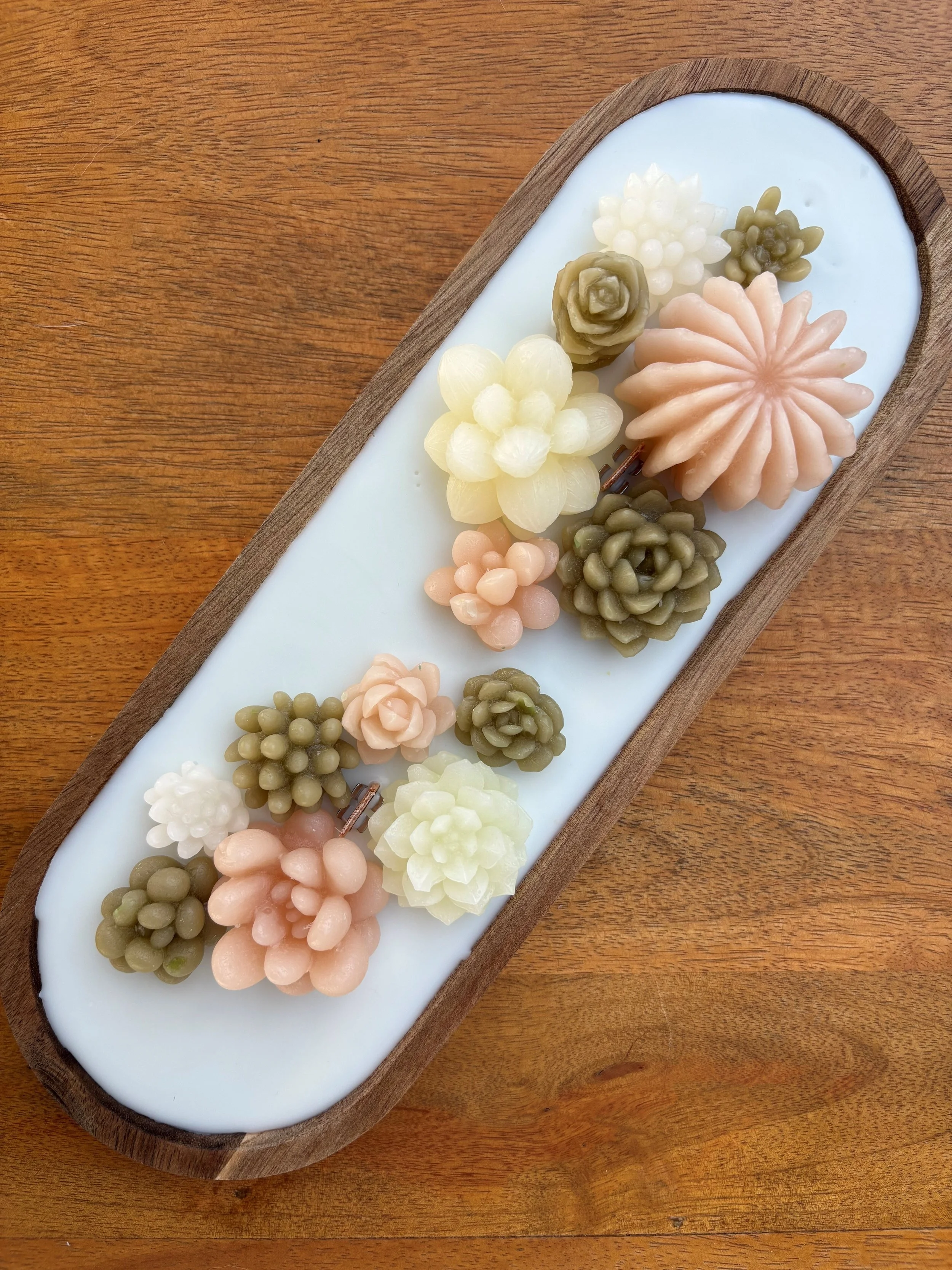 Assorted wax flowers on a white oval plate with a wooden edge, placed on a wooden surface.